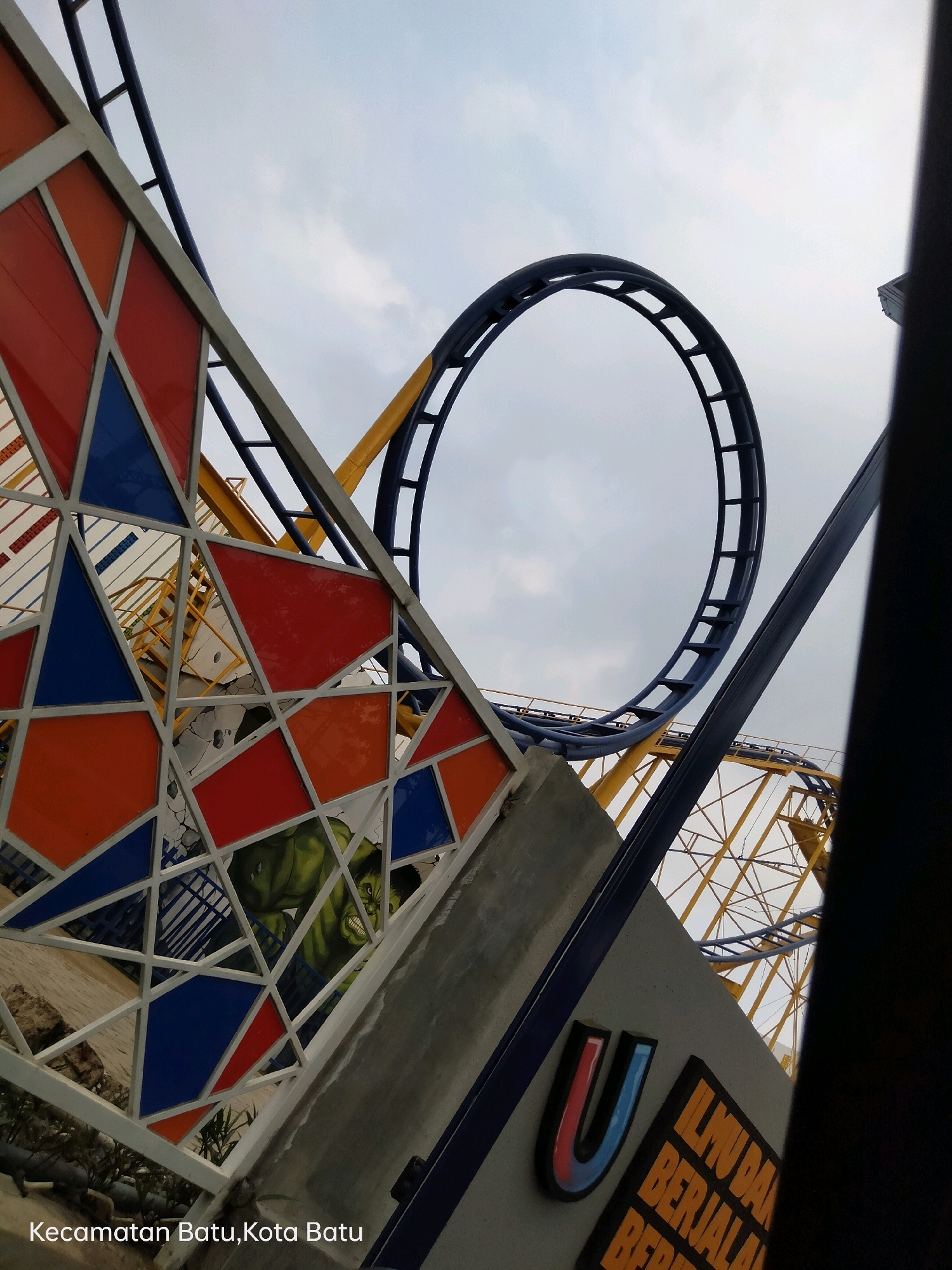 The image shows a section of a roller coaster track against a cloudy sky. The track is dark blue and forms a loop. In the foreground, there is a decorative panel with a geometric design of colored glass in red, orange, and blue. Behind this panel, a portion of the roller coaster's yellow support structure is visible, along with a mural of the Hulk. Below the mural and the decorative panel, text on a gray surface reads "Kecamatan Batu, Kota Batu," indicating the location is in Batu District, Batu City, Indonesia. A dark, vertical object, possibly part of a building or another structure, partially obscures the right side of the image.