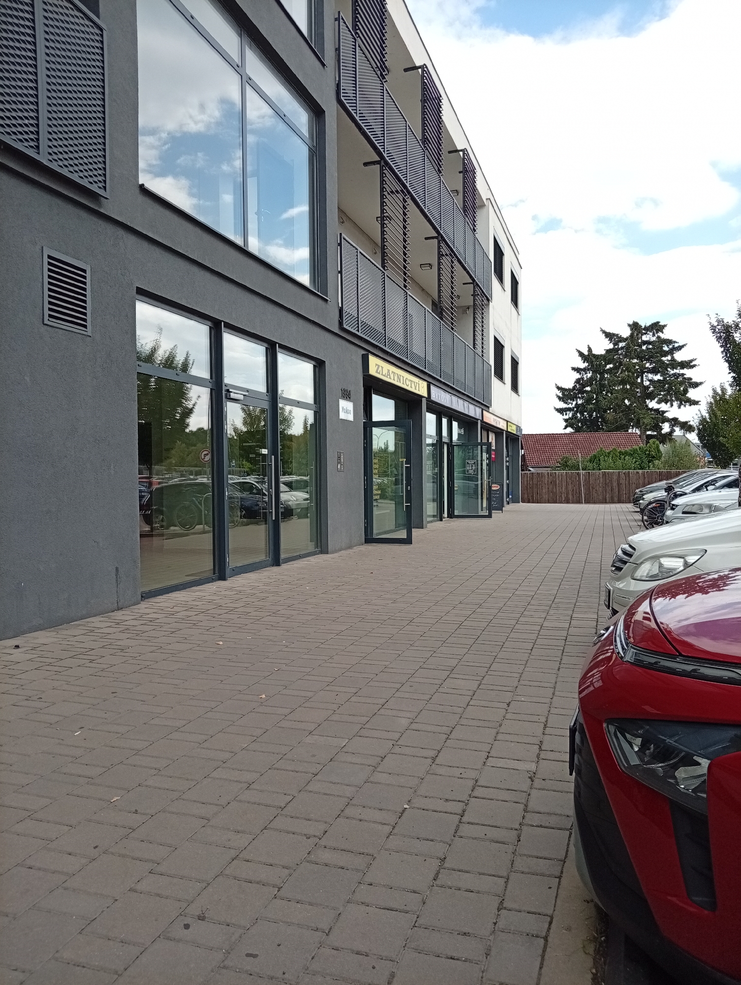The image shows a modern building with commercial spaces on the ground floor and balconies on the upper floors. The building is located on a street with a paved pedestrian walkway in front of it. Several cars are parked along the side of the street. The sky is partly cloudy. The building has large glass windows and doors, and a dark grey facade on the ground floor. Signs above the storefronts indicate businesses such as "ZLATNICTVÍ" (jewelry store). A large pine tree is visible in the background. The street appears to be in a residential or mixed-use area of Tišnov, Czechia.