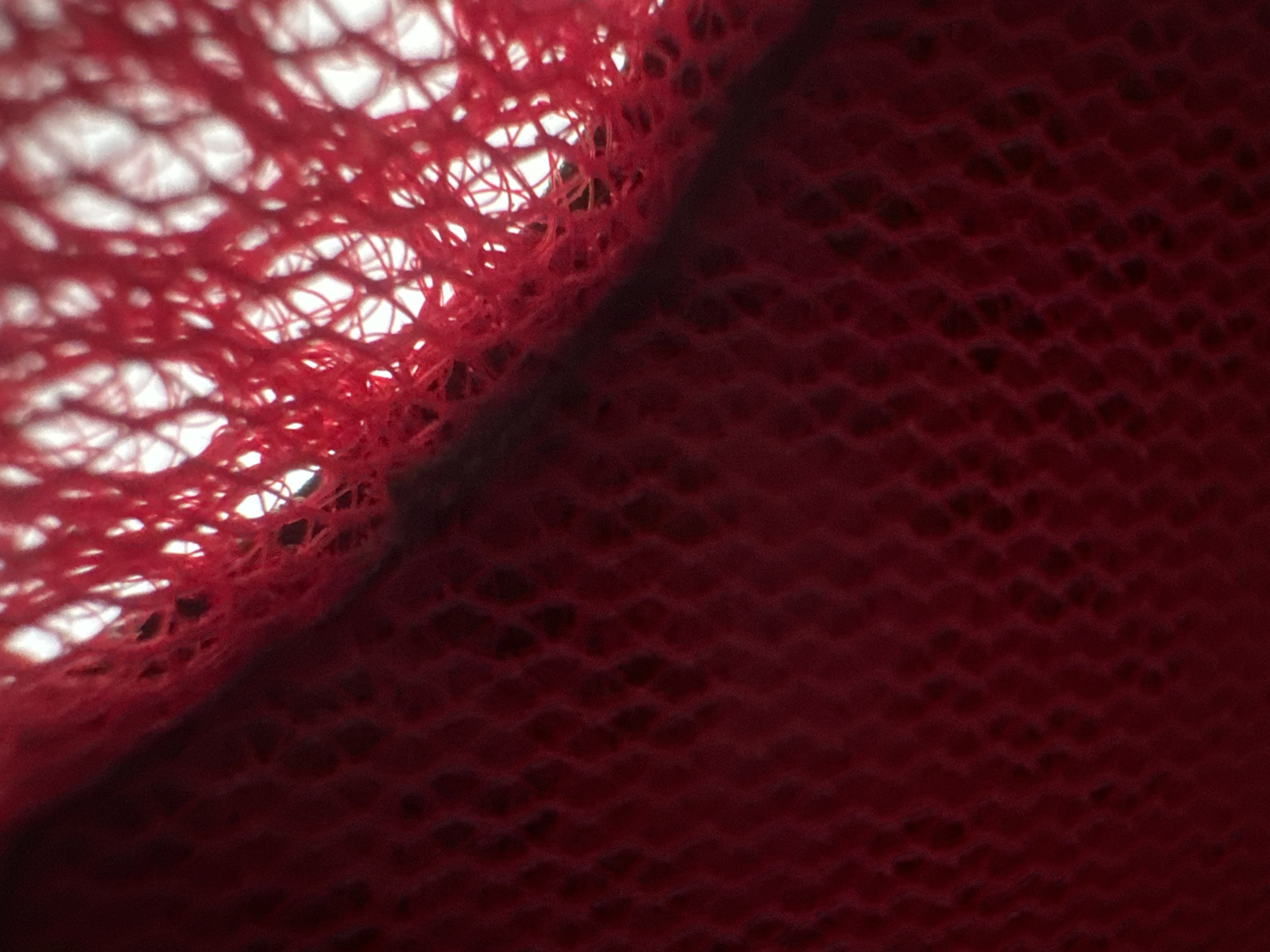 This is a close-up, abstract photograph focusing on the texture of red fabric. The image is dominated by shades of red, with a transition from a brighter, lighter red in the upper left to a deeper, darker red across the majority of the frame.
The left side of the image features a loosely woven, net-like structure in a vibrant red. Through the gaps in this netting, a bright white background is visible, suggesting that the fabric is either against a light source or is translucent. The threads of this net are somewhat frayed and irregular, giving a tactile quality to the visual.
A diagonal line divides the image, separating the net-like texture from the solid, darker red fabric on the right. This darker fabric has a subtle, textured pattern, possibly a quilted or embossed design, creating a rippled or dimpled effect across its surface. The texture here is more uniform and less open than the netting on the left.
There are no people, specific objects, or discernible scenes. The activity, if any, is not apparent from the image; it appears to be a static study of textile properties. The lighting is bright on the left, creating highlights and defining the woven structure, while the right side is more subdued, with shadows emphasizing the textural undulations.
No time of day, weather, location cues, or emotions can be inferred. There is no visible text in the image. The context of Belgrade, Serbia, does not provide any specific visual cues within this abstract close-up.