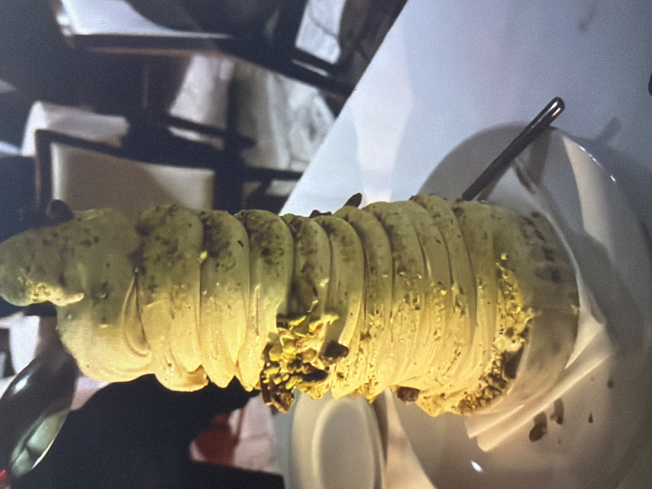 A cylindrical dessert, potentially a Kürtőskalács, rests horizontally on a white plate. The pastry appears segmented, with a light green hue and a slightly textured surface. Some segments have a visible coating of what seems to be chopped nuts, possibly pistachios. A thin metal skewer pierces through the dessert, holding it together. The dessert is positioned on a white plate, which is likely sitting on a table in a restaurant setting. The background features out-of-focus elements such as chairs and a light-colored tablecloth.