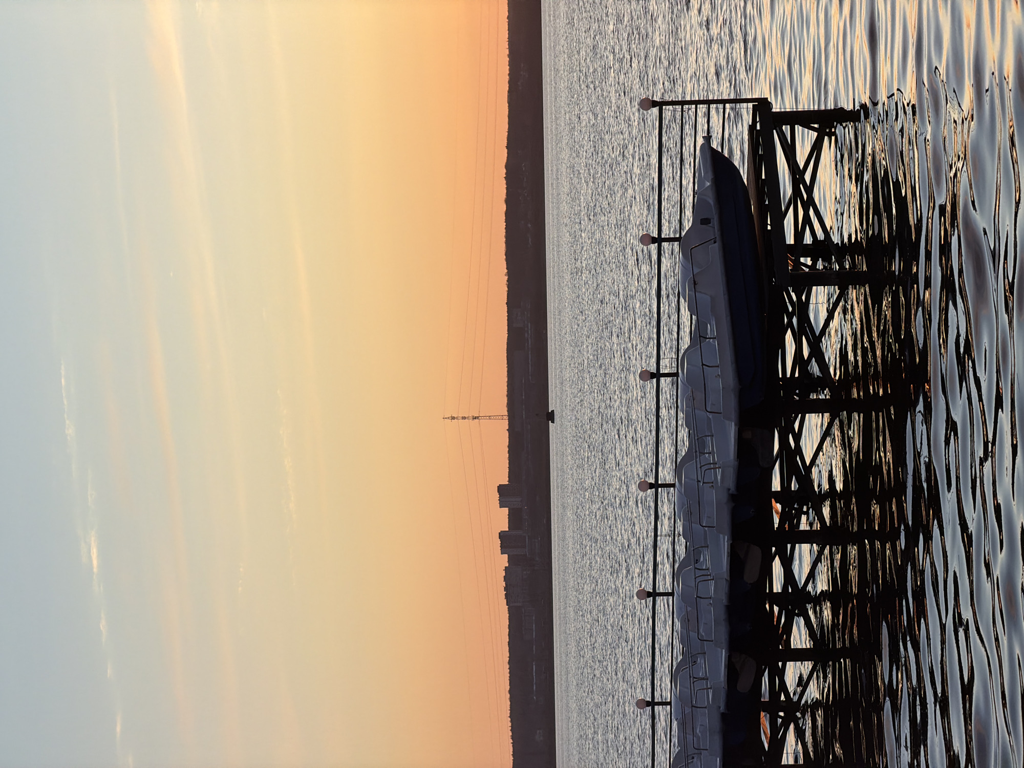 The image, captured in a portrait orientation, depicts a serene waterside scene, likely the Izhevsk Pond or Reservoir in Izhevsk, Russia, during either sunrise or sunset. The left side of the frame is dominated by the sky, displaying a warm gradient that transitions from a soft light blue and grey at the top-left to vibrant orange and yellow hues closer to the center-right horizon.
A dark, silhouetted landmass, possibly forested, stretches horizontally across the middle of the image, forming the horizon line between the sky and the water. Above this landmass, faint outlines of distant urban buildings and a tall power line tower are discernible against the glowing sky.
The right side of the image features a calm body of water, reflecting the warm colors of the sky and the dark silhouettes of objects. Gentle ripples and small waves are visible on the water's surface, creating abstract patterns of light and shadow, particularly pronounced in the foreground.
In the immediate foreground, on the right side of the frame, a dark, ornate structure, resembling a pier or dock, extends into the water. Its side features a distinctive criss-cross or lattice-like pattern. Lined up neatly along this dock are several white or light-colored pedal boats, currently idle and resting on the water. Three spherical, white lights are mounted on vertical posts along the railing of the dock.
No people or active events are visible in the tranquil scene. The overall mood is peaceful and calm, capturing a quiet moment at dusk or dawn over the water. No visible text is present.