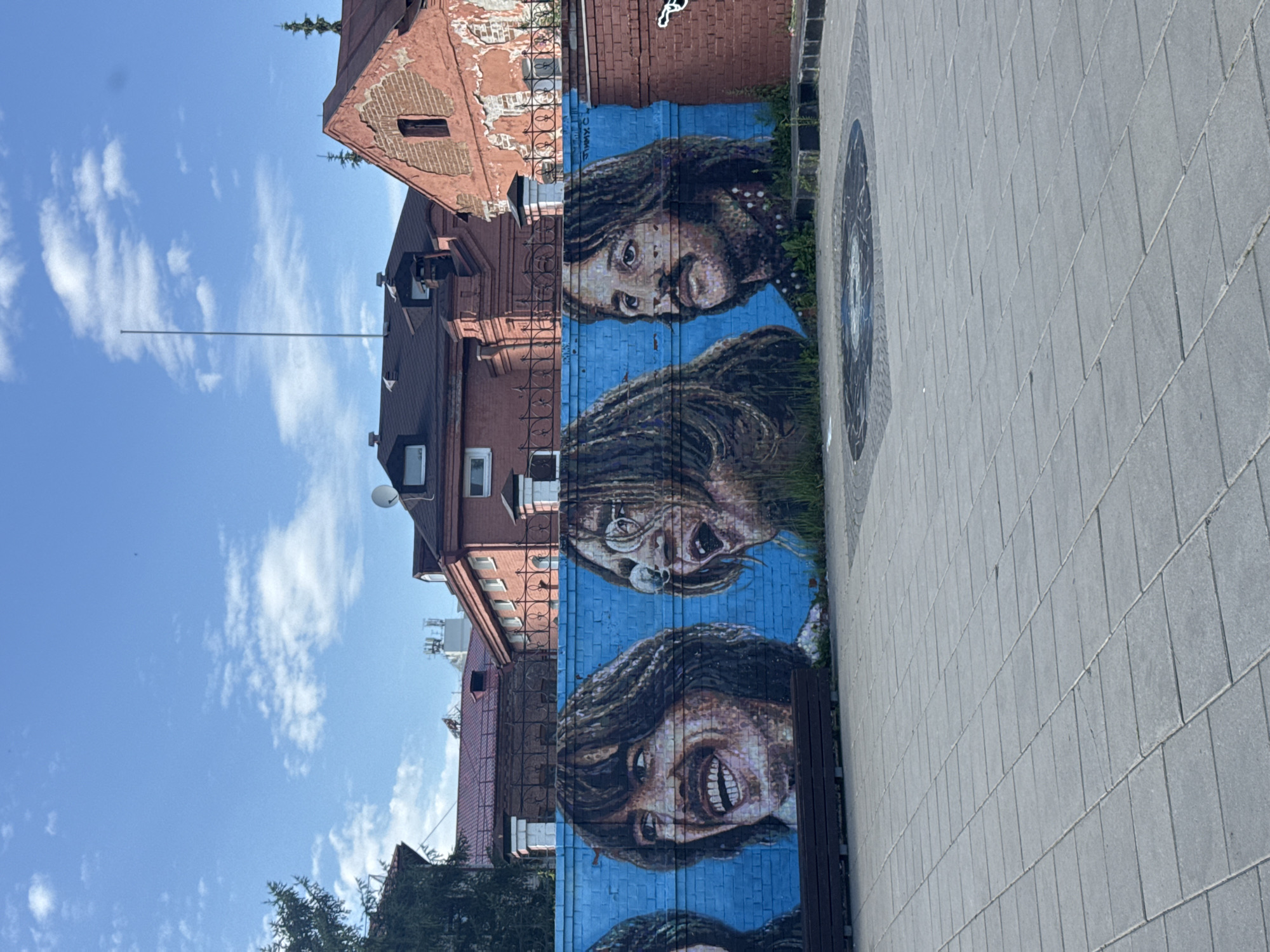 The image depicts a mural painted on the side of a brick building in Yekaterinburg, Russia. The mural features three large portraits of people with striking expressions, their faces rendered in a colorful and detailed style against a vibrant blue background. The subjects appear to be in mid-expression, with open mouths and wide eyes, conveying a sense of surprise or excitement. The architectural style of the building, with its red brickwork and visible windows, suggests an urban environment. Adjacent to the mural is a paved walkway made of grey rectangular stones, and a dark wooden bench is situated in front of the artwork, inviting viewers to pause and admire it. The scene is set against a bright blue sky with scattered white clouds, indicating fair weather and likely daytime. The overall impression is one of public art in an urban setting, adding a lively and artistic element to the streetscape. No visible text is present in the image, apart from what might be artist signatures or titles, which are too small to discern clearly.