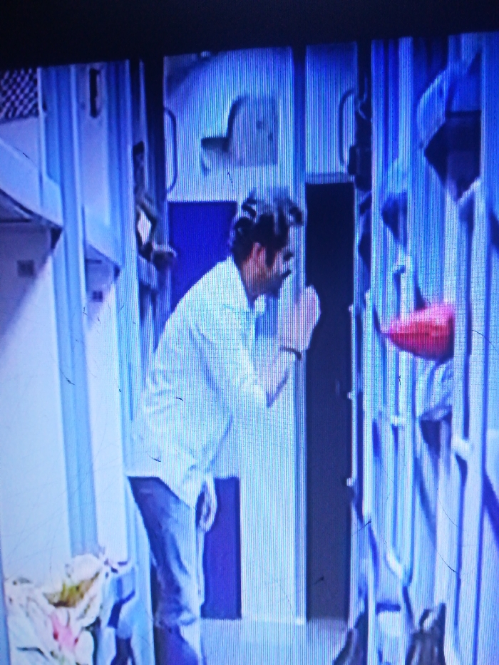 A man in a white shirt and jeans stands in a hallway with bunk beds on the right. He appears to be looking at something or someone just out of frame. The hallway is painted in shades of blue. There are items stored near the lower bunk on the left side, and a red object is visible on the bunk beds on the right. The scene looks like it could be inside a train or a similar space with confined sleeping quarters.