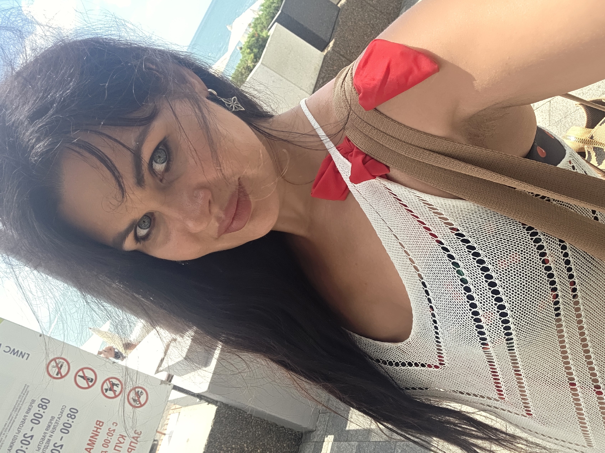A medium shot captures a woman with long, dark hair styled loosely, looking directly at the camera. She has fair skin and striking blue-green eyes. Her mouth is slightly open, and she is wearing a white, open-knit top with a patterned garment underneath. Two red bow-shaped adornments are attached to the straps of her top, one on her left shoulder and another on the strap crossing her chest. A brown strap from a bag is visible across her right shoulder. She is outdoors, with a blurred background suggesting a sunny day with a body of water and some greenery. To the left of the frame, a sign with various prohibition symbols and text in Russian is partially visible. The overall impression is a casual, candid portrait taken in a public outdoor setting, likely a resort area. The signage on the left, specifically the text "LWWC", and the overall aesthetic of the location are consistent with coastal areas in Sochi, Russia.