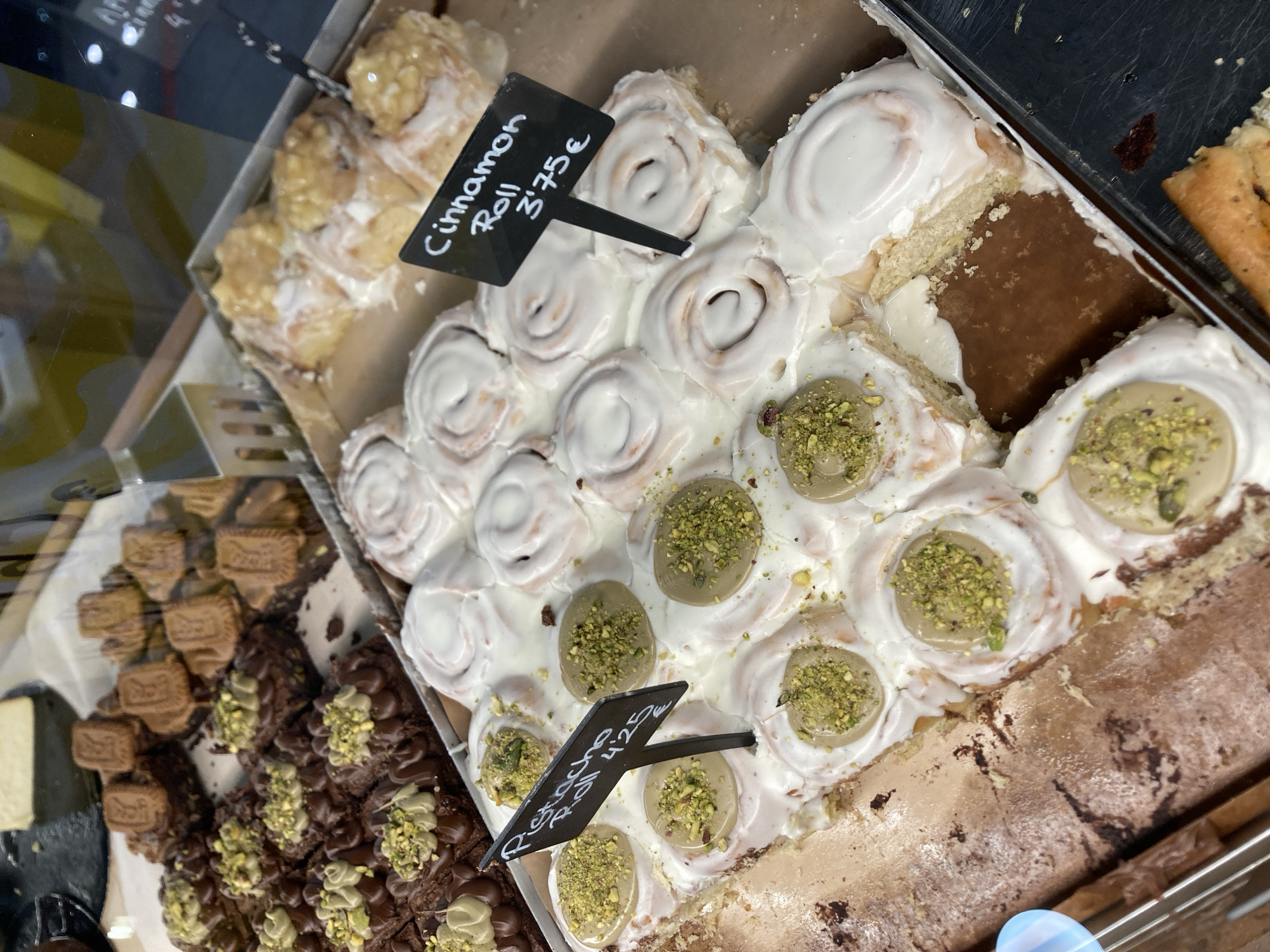 The image shows a close-up view of a display of pastries in a bakery or pastry shop. The pastries appear to be arranged in trays, suggesting they are for sale.
The primary subjects are various types of baked goods. In the foreground and center, there are numerous cinnamon rolls covered in white icing. Some of these cinnamon rolls are topped with a dollop of light green cream and sprinkled with chopped pistachios. To the left of the cinnamon rolls, there are dark chocolate brownies, also topped with a similar green cream and pistachios. Further to the left, there are small, rectangular chocolate biscuits. In the background, on the left, there is a tray of what appear to be lighter colored pastries, possibly also cinnamon rolls or sweet buns, with a glossy glaze.
A black price tag is visible in the center of the image, partially obscuring some of the cinnamon rolls. The visible text on this tag reads "Cinnamon Roll 3.75€". Another similar price tag is visible further down, reading "Pistacho Roll 4.25€". The pastries look fresh and appealing, indicative of a food retail environment.
The setting is indoors, likely a bakery or cafe, as suggested by the display trays and the variety of baked goods. There are no people visible in the frame. The lighting appears to be artificial, typical of an indoor commercial space. The overall impression is one of a tempting display of sweet treats. The location context of Barcelona, Spain, might suggest the style or ingredients of the pastries, but this cannot be definitively determined from the visual information alone.