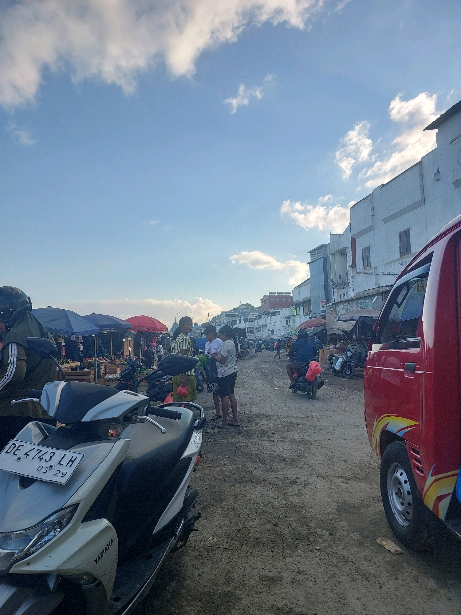 The image depicts a street scene in Ambon, Indonesia, likely a marketplace given the stalls and goods visible. The sky is blue with scattered clouds. In the foreground, a white and silver Yamaha scooter is parked, its license plate reading "DE 4743 LH 03 29". To the right, the side of a red vehicle, possibly a small truck or van, is visible.
The street is unpaved and dusty, with several people present. Some individuals are standing near market stalls selling produce, indicated by wooden crates filled with fruits and vegetables. Others are on or near motorcycles. In the mid-ground, a person wearing a blue jacket is riding a motorcycle away from the camera. Further down the street, buildings line the road, some appearing to be shops or residences. Umbrellas, some red and some blue, are set up to provide shade for the market stalls. The overall impression is that of a busy, casual market environment in a tropical or subtropical climate.