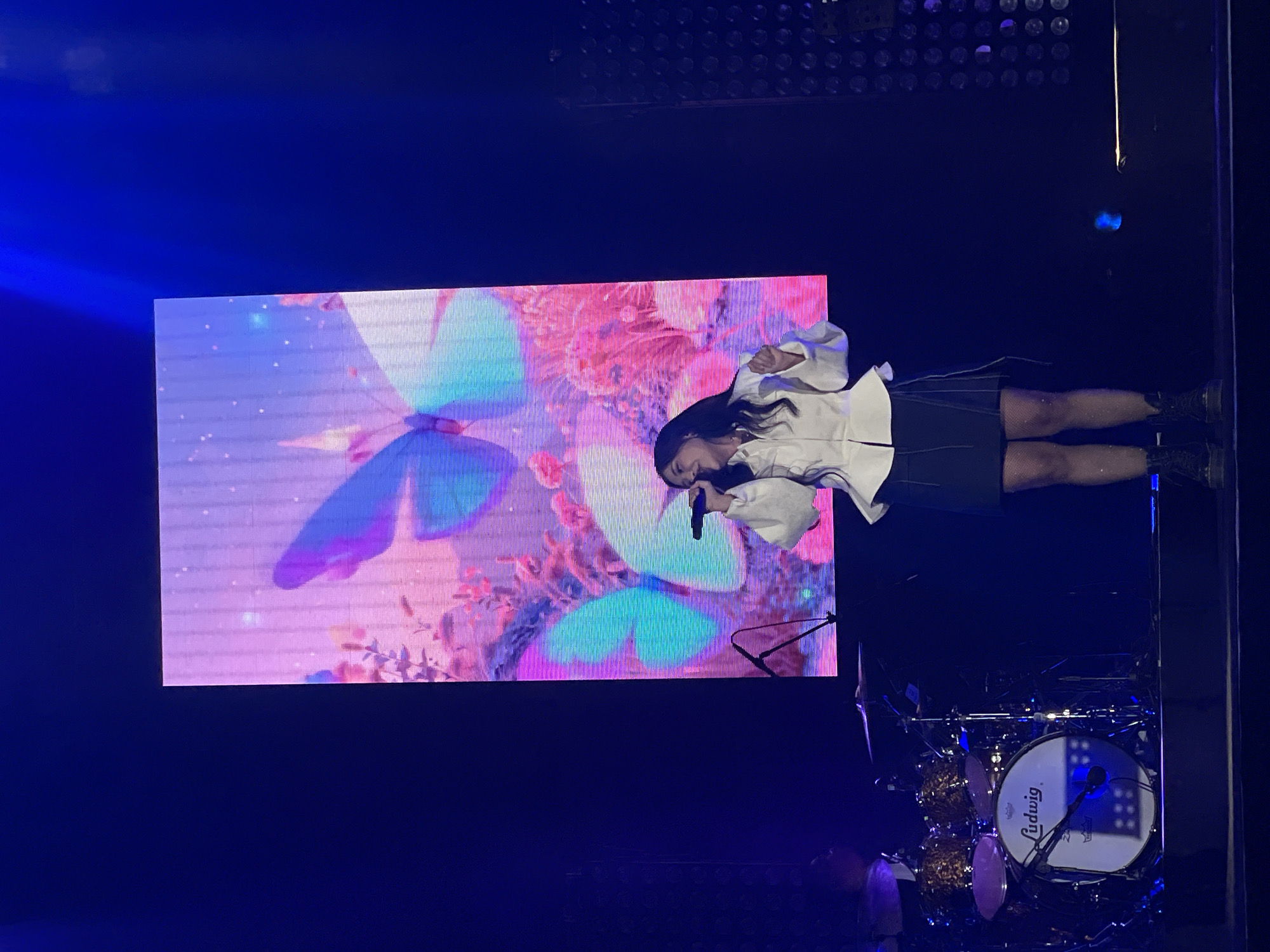 A woman in a white, long-sleeved shirt and black skirt stands on a stage, singing into a microphone. Behind her is a large screen displaying a vibrant image of butterflies and flowers. To the right, a Ludwig drum set is visible. The location appears to be a concert setting, possibly in Salembaran, Indonesia.