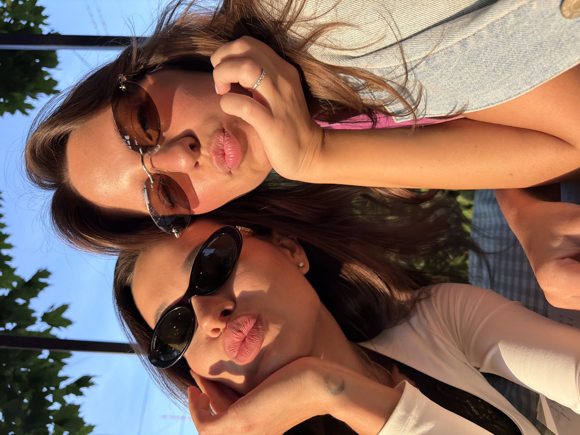 Two young women, each wearing sunglasses and making kissing faces, are captured in a close-up, selfie-style photograph. The woman on the left has dark hair and is resting her chin on her hand. She is wearing a white top. The woman on the right has lighter brown hair and is also resting her chin on her hand. She is wearing a pink top and a denim jacket. Both women appear to be outdoors, with dappled sunlight illuminating their faces. In the background, green foliage and a blue sky are visible, suggesting an outdoor setting such as a park or a restaurant with outdoor seating. The presence of what appears to be a checkered tablecloth and some papers suggests they might be at a cafe or dining outdoors. No specific landmarks in Saint Petersburg, Russia are identifiable in this image.