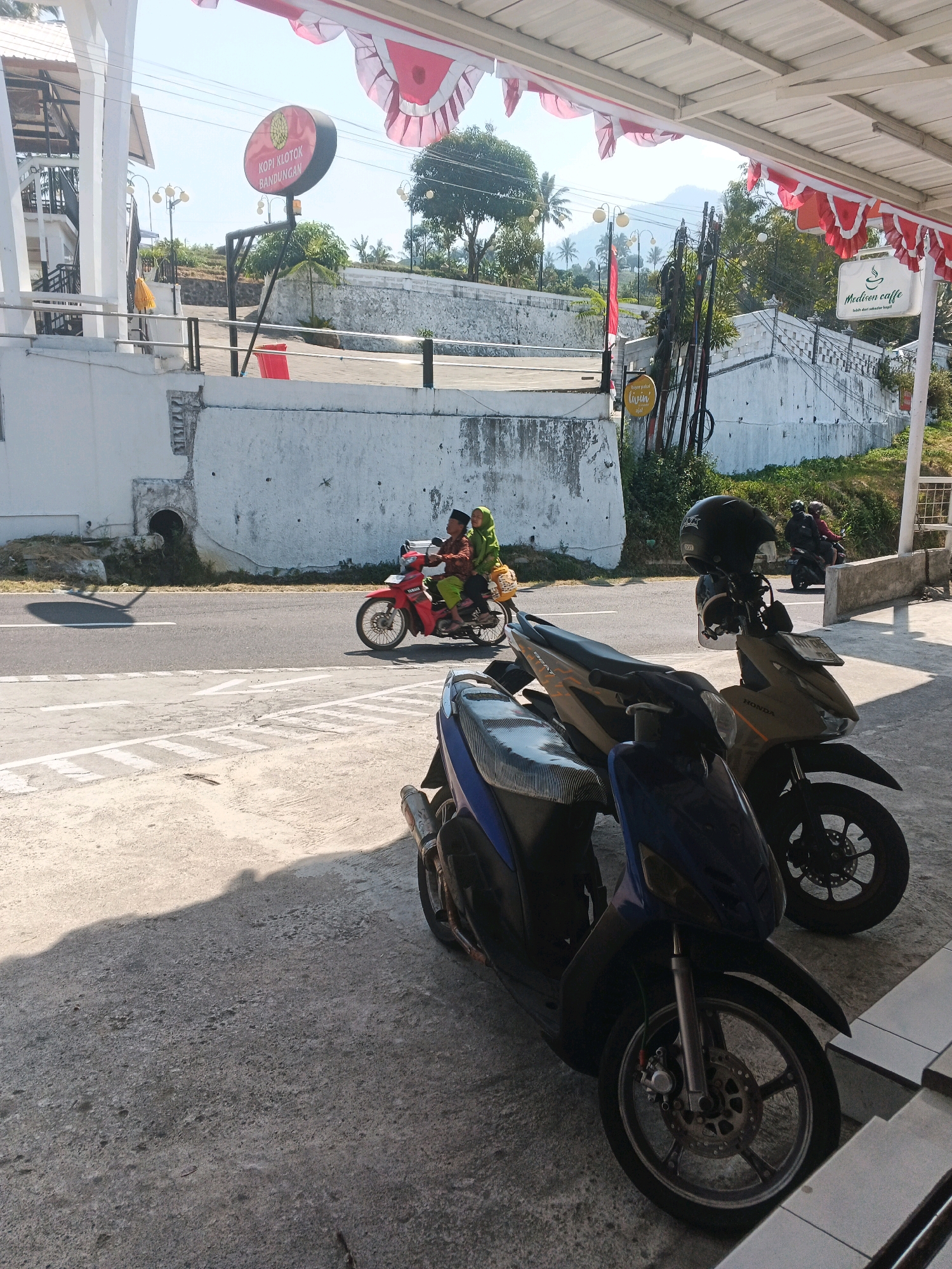 In Pakopen, Indonesia, a street scene unfolds under a clear, bright sky. In the foreground, two parked scooters, one blue and one tan, sit on a paved surface. The blue scooter is closer, its front wheel and handlebars clearly visible. Behind it, the tan scooter has a black helmet resting on its handlebars. Beyond the scooters, a red motorcycle travels on a road, carrying two passengers dressed in traditional clothing. The road is bordered by a white wall, behind which lush greenery and trees are visible. Several signs are posted along the road, including a circular sign with red and yellow lettering that reads "KOPI KLOTOK BANDUNGAN" and a white sign with green lettering that says "Mediven caffe". Red and white banners, typical of Indonesian flags, are strung across the top of the frame, suggesting a festive occasion or patriotic display. The overall impression is one of a vibrant, bustling Indonesian town on a sunny day.