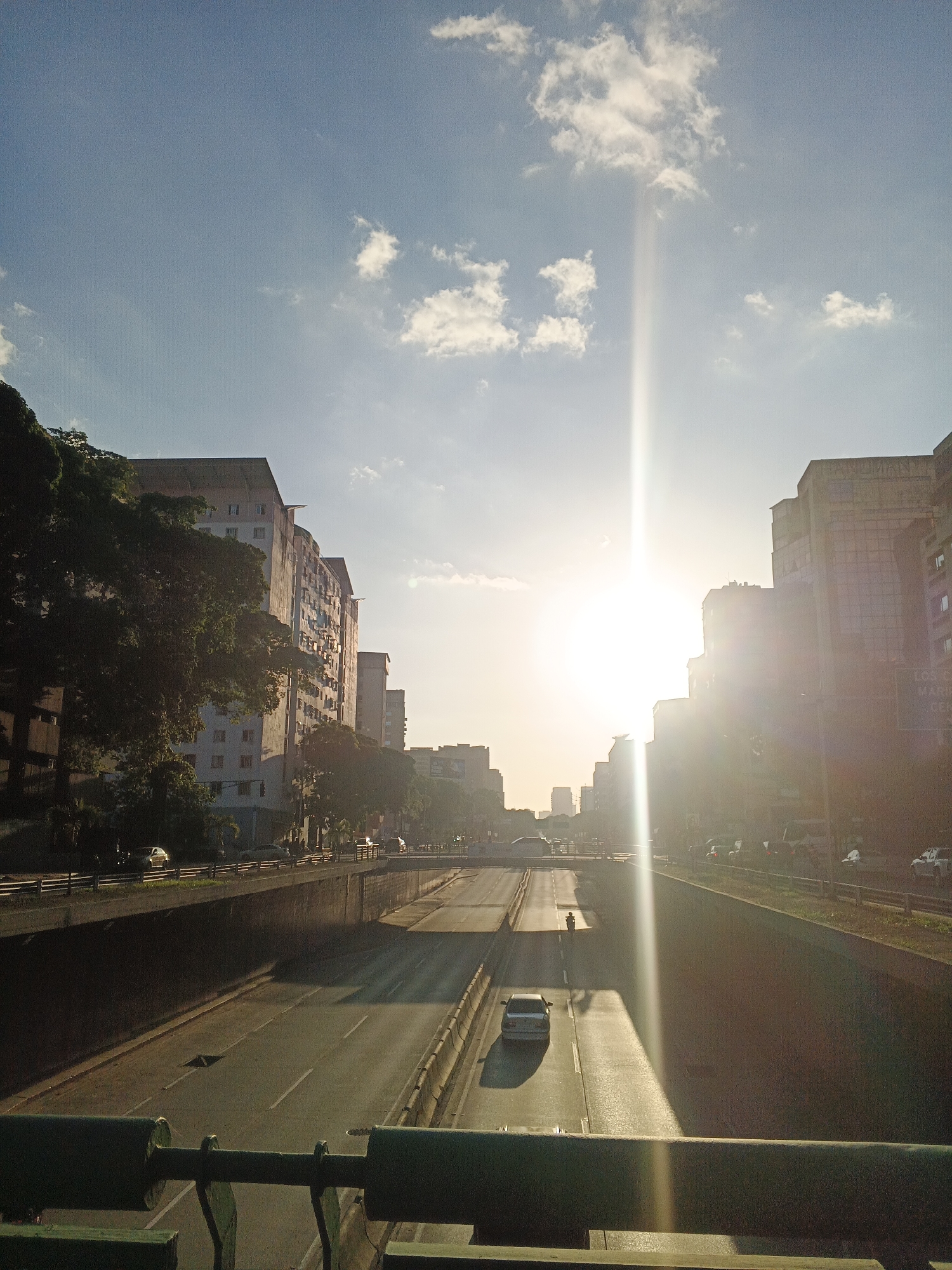 This image depicts a view of a highway in Caracas, Venezuela, looking towards the sun during a late afternoon or early morning. The highway, partially cut into the landscape, has multiple lanes of traffic with several cars visible. On the left side of the highway, large apartment buildings are prominent, partially obscured by lush green trees. On the right side, taller modern buildings line the route, with some signage visible on them. The sun is very bright, creating a strong lens flare that extends vertically through the image, casting long shadows and highlighting the textures of the road and surrounding structures. The sky is a clear blue with a few wispy clouds. The overall impression is an urban scene bathed in the warm light of the sun.