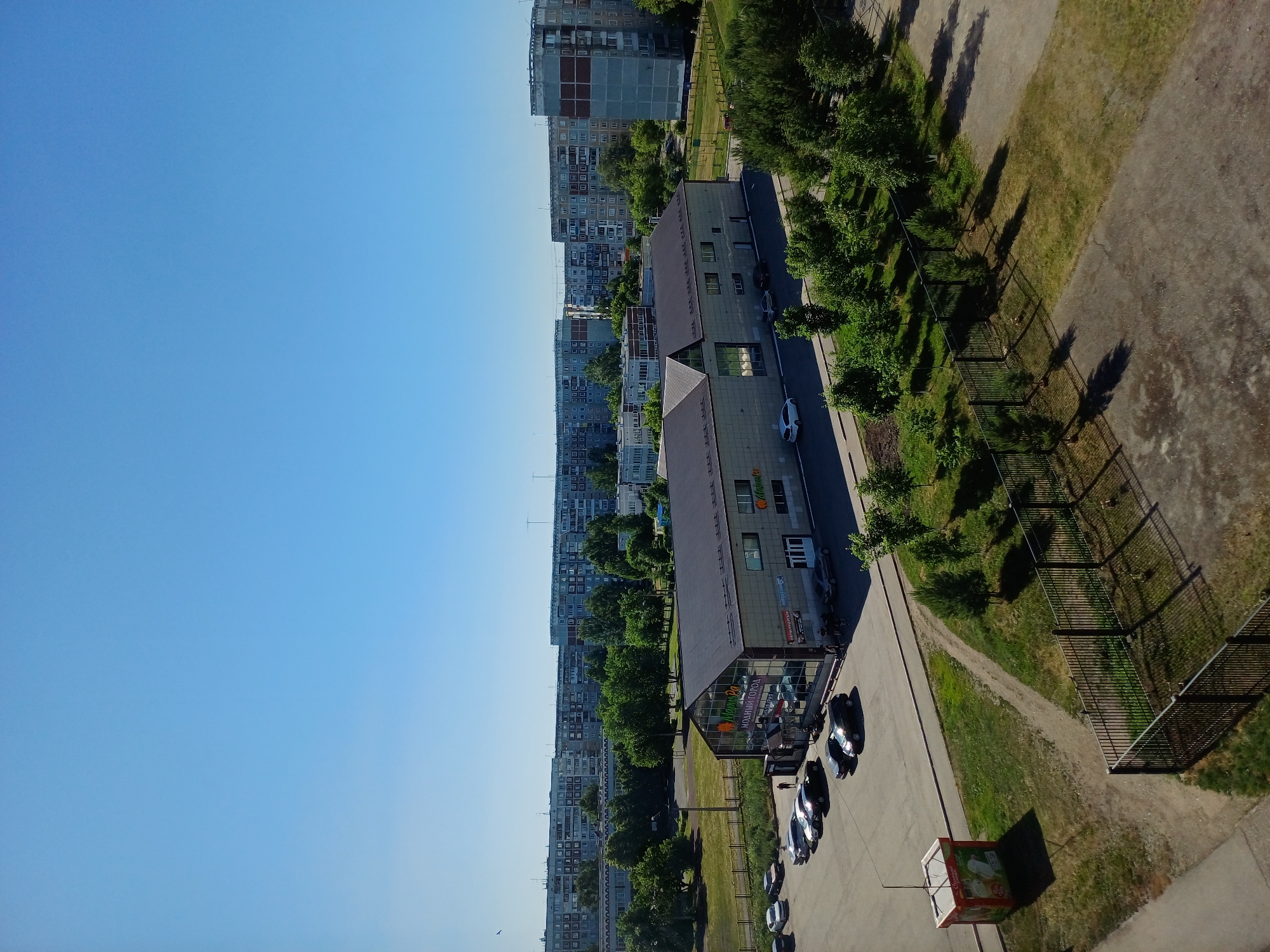 The image depicts a clear, sunny day in an urban environment. In the foreground, a black fence encloses a grassy area with scattered trees and long shadows cast by the sun. Adjacent to this, a parking lot is visible with several cars parked. A small, red and white kiosk stands on the left side of the parking lot.
Further back, a long, low-rise building with a dark roof and a facade made of light-colored rectangular tiles is prominent. This building has large glass windows and signs, including one that appears to read "МОДНЫЙ ГОРОД". A few people are visible near the entrance of this building.
Beyond the low-rise building, a dense line of green trees separates it from a series of multi-story residential buildings. These apartment blocks are typical of Soviet-era architecture, with uniformly structured facades and many windows. The sky above is a clear, bright blue, indicating good weather. The overall scene suggests a residential neighborhood on a summer day.
