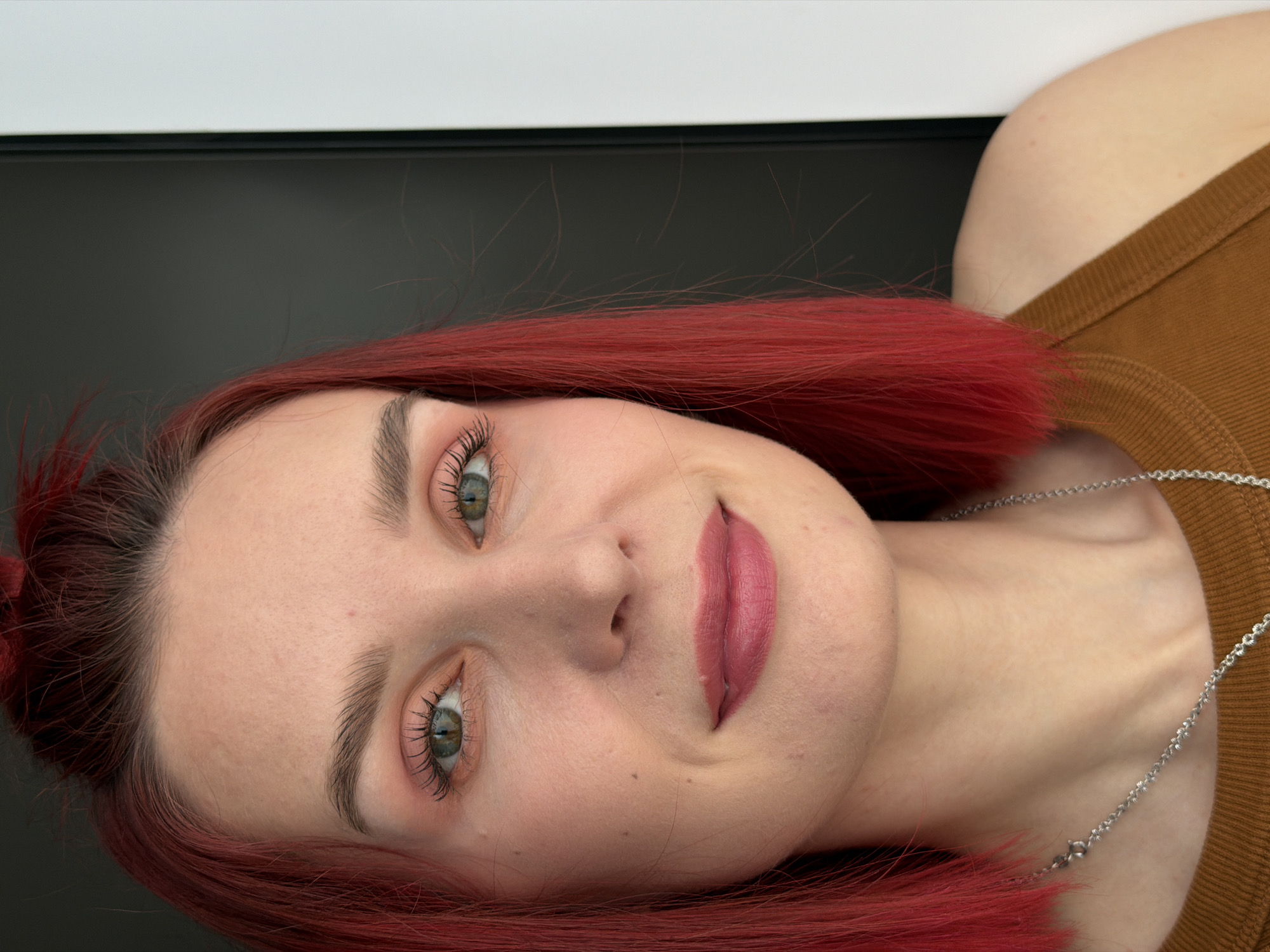 This close-up portrait, taken in Chaikovsky, Russia, features a woman with vibrant red hair, oriented horizontally as if lying down or with her head tilted sharply to the side. Her head and upper body fill the frame, set against a simple, uncluttered background that transitions from a dark grey/black to a lighter white strip at the top.
The subject is a woman with fair skin, prominent green eyes, and distinct dark eyebrows. Her eyelashes are long and full, suggesting the use of mascara or extensions. Her lips are painted with a matte, pinkish-mauve lipstick, and she has a subtle, closed-mouth smile or smirk. A faint dimple or natural indentation is visible on her chin. Her red hair, which appears to be a solid, rich color, frames her face and spreads around her head.
She is wearing a textured, ribbed brown garment, likely a top, and a delicate silver chain necklace is visible around her neck, resting on her collarbone area. The lighting is soft and even, highlighting her facial features without harsh shadows, and there are no discernible external location cues beyond the provided context of Chaikovsky, Russia, indicating an indoor setting. The woman's expression is calm and direct, looking towards the viewer. There is no visible text in the image.