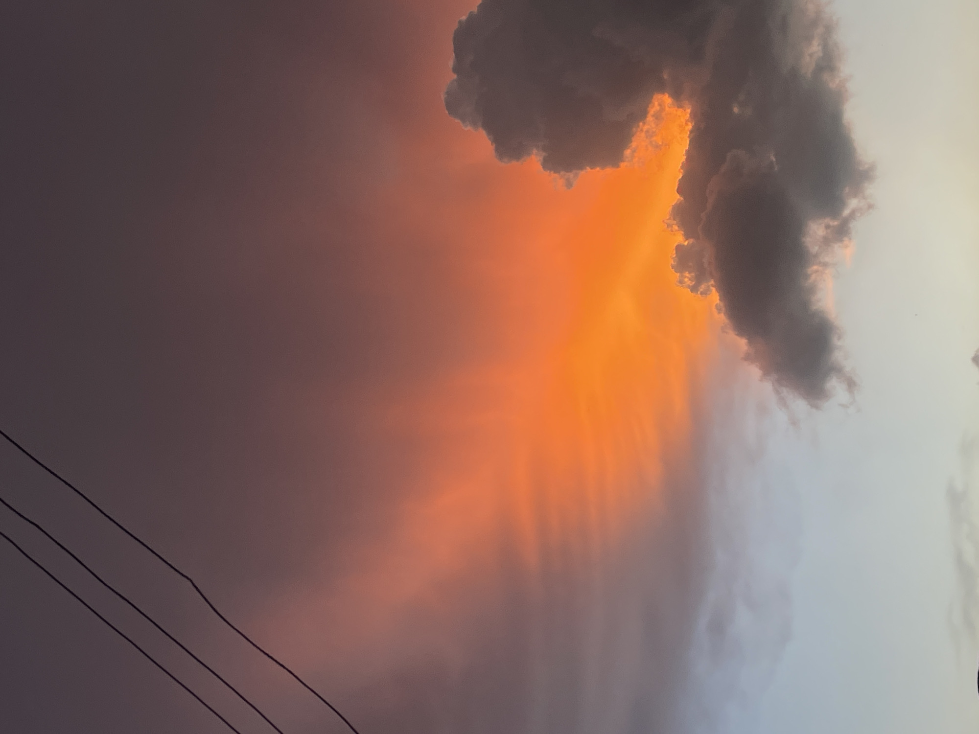 A vertical, eye-level photograph captures a dramatic sunset over the city of Jos, Nigeria. The sky is filled with vibrant hues of orange and pink, transitioning to a dusky purple at the top of the frame. The sun, not directly visible, illuminates the clouds from behind, creating a fiery glow that radiates outwards. A large, dark cloud formation sits on the right side of the image, its edges illuminated by the setting sun. Three dark, thin power lines diagonally traverse the upper left quadrant of the sky. The lower portion of the image shows a lighter, pale blue sky with faint clouds. The scene is devoid of any people, specific landmarks, or identifiable structures within the frame, suggesting a focus solely on the natural spectacle of the sunset.