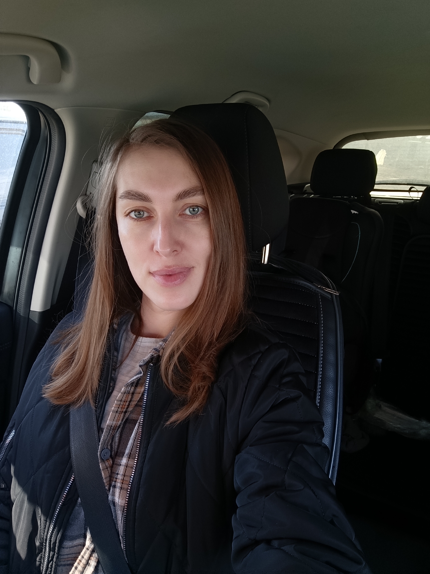 A medium shot depicts a young woman seated in the driver's seat of a car, wearing a black quilted jacket over a plaid shirt. She has long, light brown hair and light-colored eyes. She is wearing a seatbelt. The car's interior is visible, including the front seats and headrests. Based on visual information alone, no information suggests this scene is specifically located in the city of Chaikovsky, Russia.