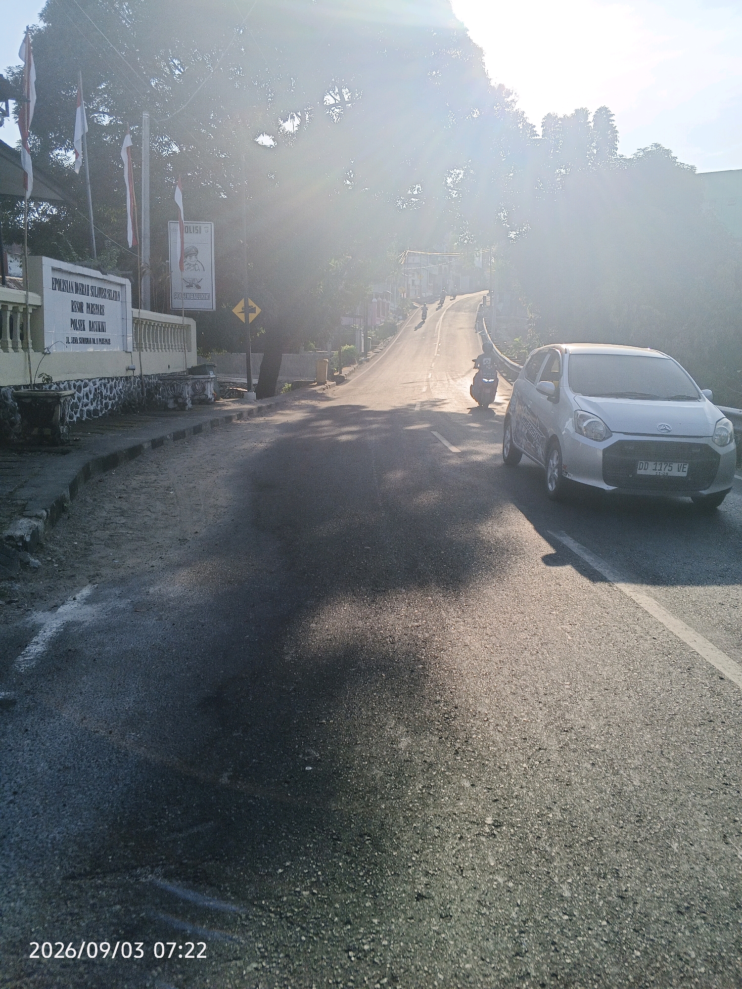 The image depicts a street scene in Pare-Pare, Indonesia, bathed in the warm light of what appears to be sunrise or sunset. On the left side of the road, there is a low, cream-colored building with a stone foundation and a decorative railing. Above the building, several Indonesian flags are displayed on poles, fluttering in the breeze. A sign on the building reads "EPOLISTAS DAERAH SULAWESI SELATAN RESOR PAREPARE POLSEK BACUKIKI", indicating it is the Polsek Bacukiki (Bacukiki Police Station) within the Pare-Pare Resort of South Sulawesi Regional Police.
The road itself is asphalted and appears to be slightly damp in places, with some white markings. A silver car is parked or driving on the right side of the road, facing away from the viewer. In the distance, the road curves uphill and is lined with trees and other vegetation. Several motorcycles are visible on the road, suggesting ongoing traffic. The overall atmosphere is serene, with the low sun casting long shadows and creating a lens flare effect. The timestamp at the bottom left of the image indicates the date and time as 2026/09/03 07:22.