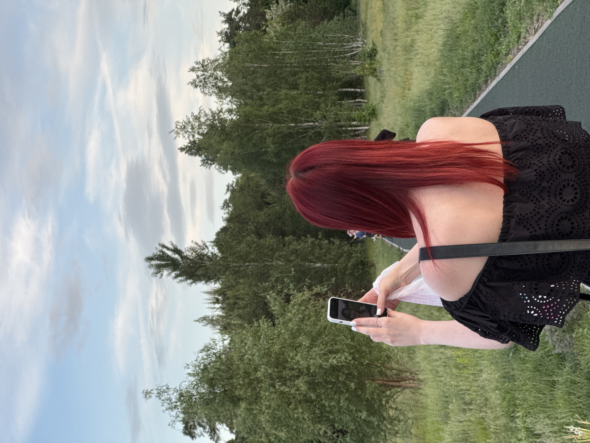 A woman with auburn hair and wearing a black blouse is photographed in Unknown, Unknown. She is holding a white smartphone in her hands. The background features trees and grass, with a path visible on the right side of the frame. The sky is visible on the left side, with clouds.