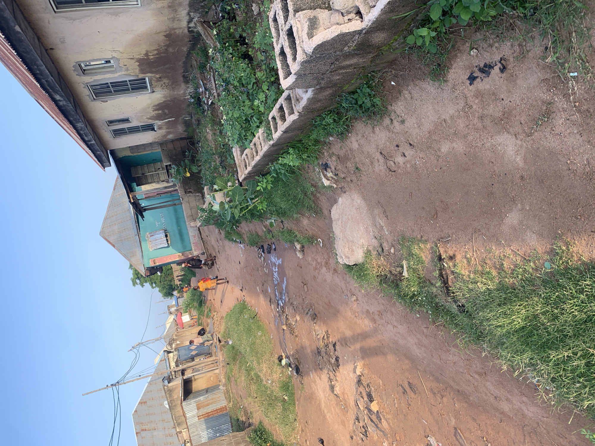 The image depicts a dirt road in a residential area, likely in Jalingo, Nigeria, given the context. The scene is characterized by a slightly elevated perspective, looking down a winding, unpaved road. The ground is a reddish-brown dirt, with evidence of scattered debris, including dark, wet-looking patches and some white material. There are patches of dry, sparse grass growing along the sides of the road and on the verges.
On the right side of the road, a low wall constructed from concrete blocks is visible, with green vegetation growing behind it. Further into the background, several structures are present. On the far left, a building with a corrugated metal roof is visible, appearing somewhat dilapidated. Adjacent to it, another building with a similar roof and corrugated metal siding is seen.
Further down the road, a teal-colored building stands out, featuring windows and a sign that is partially legible, possibly indicating a "GREEN HOUSE." A man in a black garment and a young boy in an orange t-shirt are walking away from the viewer on the road. Another individual, carrying a dark bag, is walking in the same direction further up the road. The sky above is a clear, pale blue, suggesting it is daytime. The overall impression is of a humble, possibly developing, urban environment with signs of everyday life and informal construction.