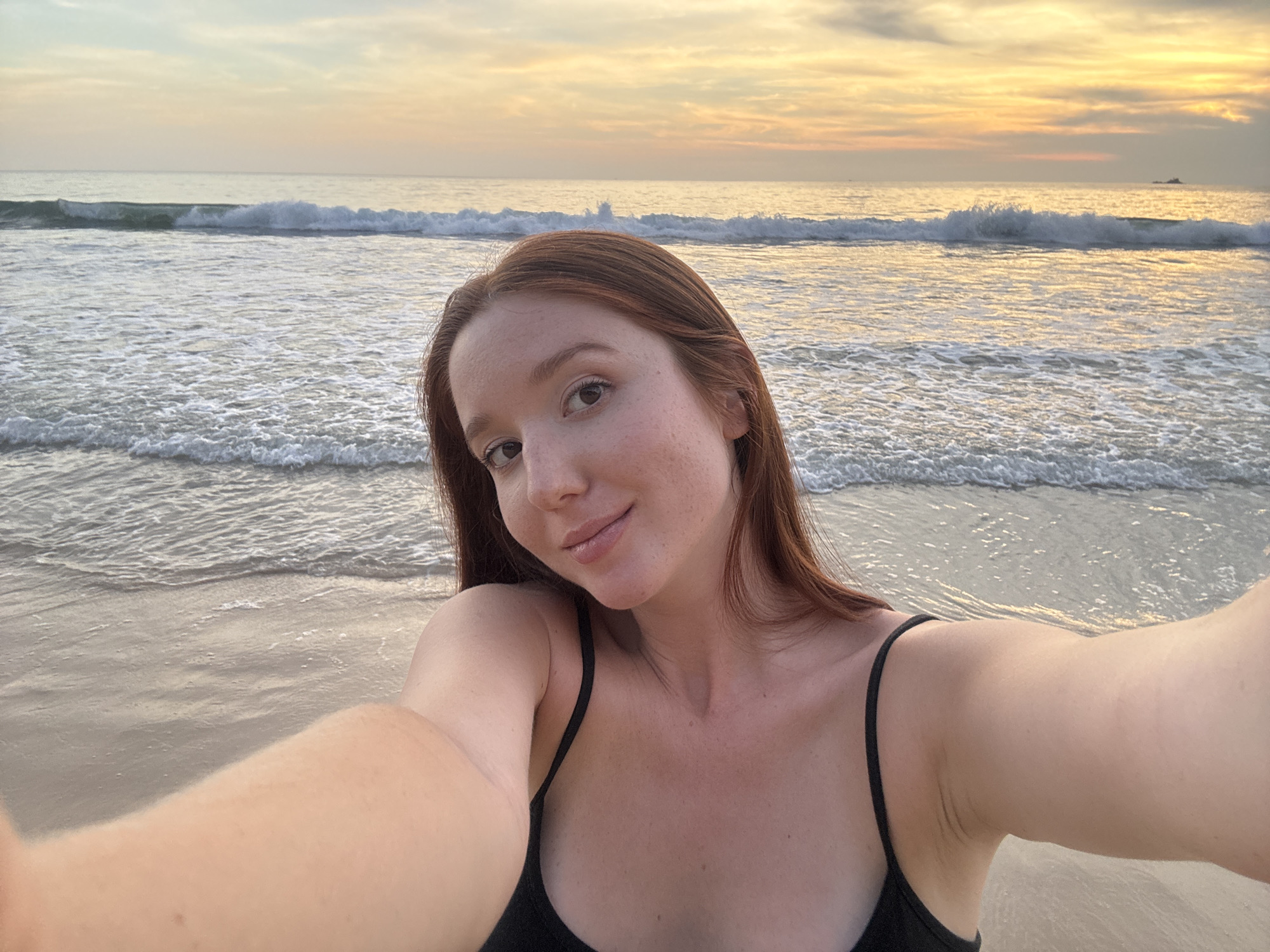 A woman takes a selfie at sunset on Bang Tao Beach in Choeng Thale, Thailand. The beach is sandy and wet, with small waves in the background. The sunset creates an orange glow on the water and sky. The woman is wearing a black top. Her arm is extended to hold the camera.