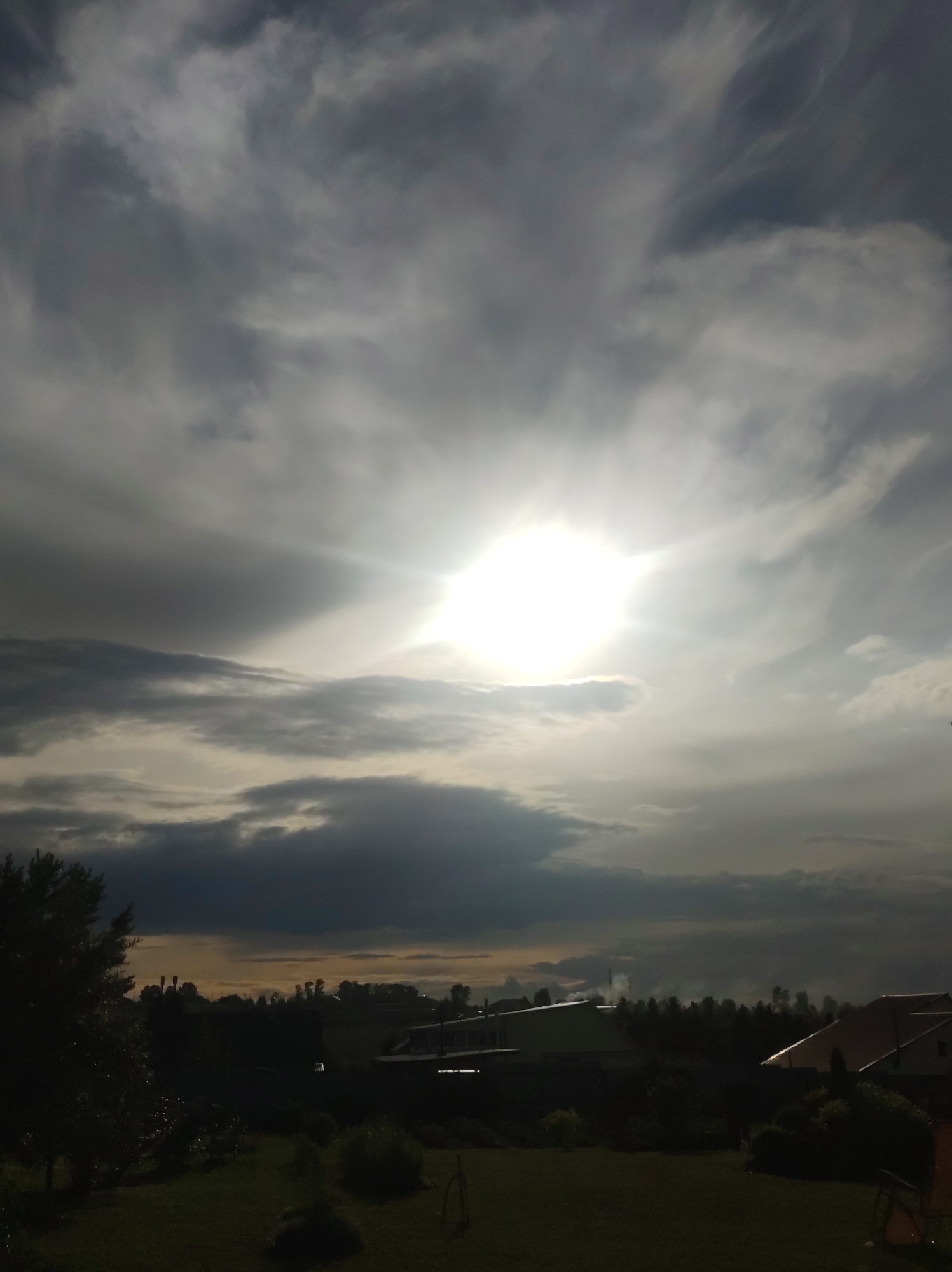 The image depicts a cloudy sky with the sun shining brightly through the clouds, creating a sunburst effect. Below the sky, a silhouetted landscape of trees and houses is visible, suggesting a rural or suburban setting. The lighting indicates it is likely sunset or sunrise, as the sun is low in the sky and the clouds are illuminated. The overall mood is serene and perhaps slightly dramatic due to the contrast between the bright sun and the dark clouds and landscape. There is no visible text. The location is identified as Фоки, Russia. The scene appears to be a peaceful, natural setting with human habitation in the distance.