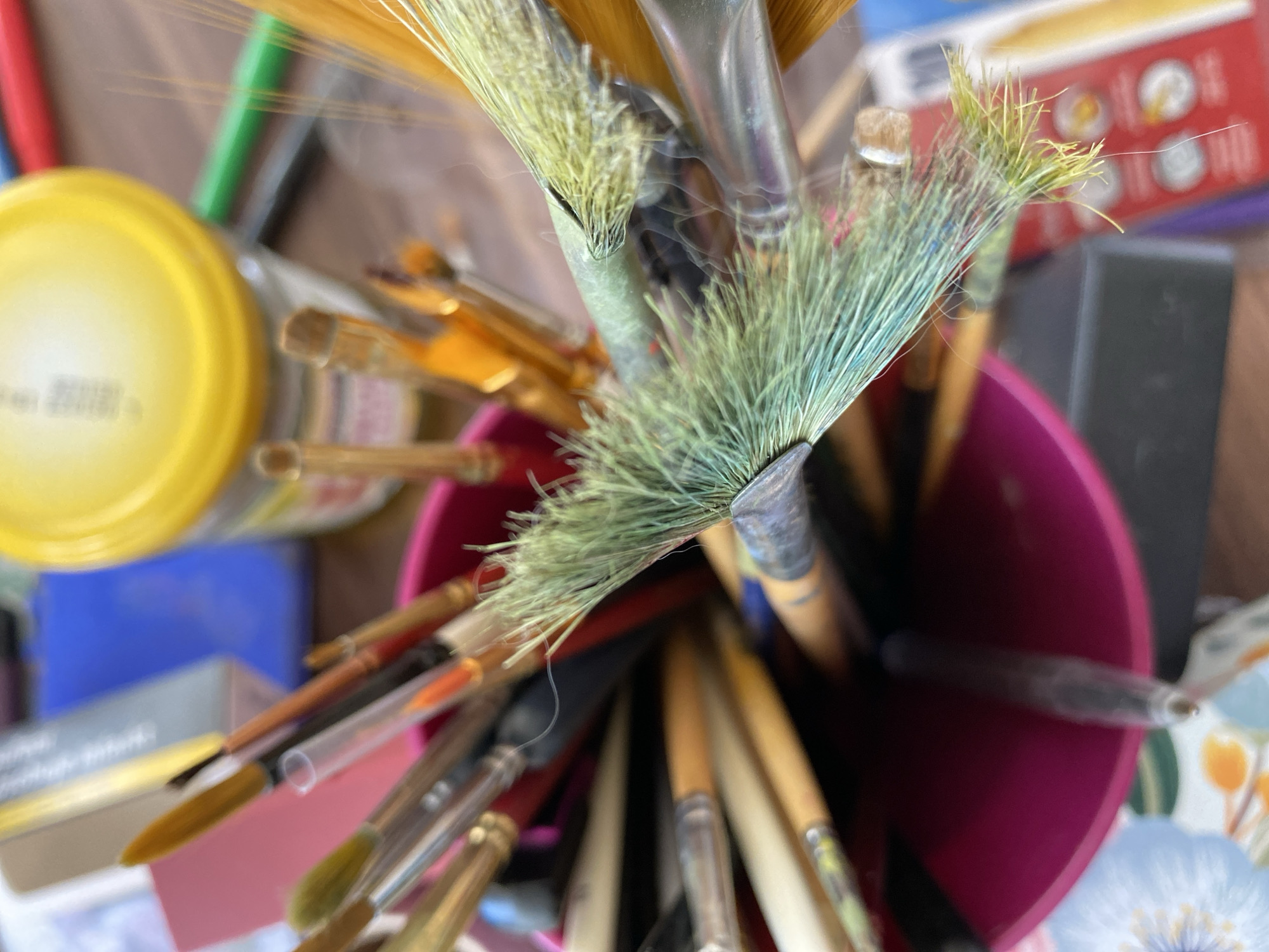 The image shows a close-up, top-down view of a collection of art brushes held in a pink container. The brushes have varying bristle colors and sizes, including green, yellow, and brown. Some brushes appear to be watercolor or acrylic brushes, with fine bristles. In the background, out of focus, are art supplies such as a yellow lidded jar, colored markers, and a box. The overall impression is of an artist's workspace. The image was taken in Bílina, Czechia.
