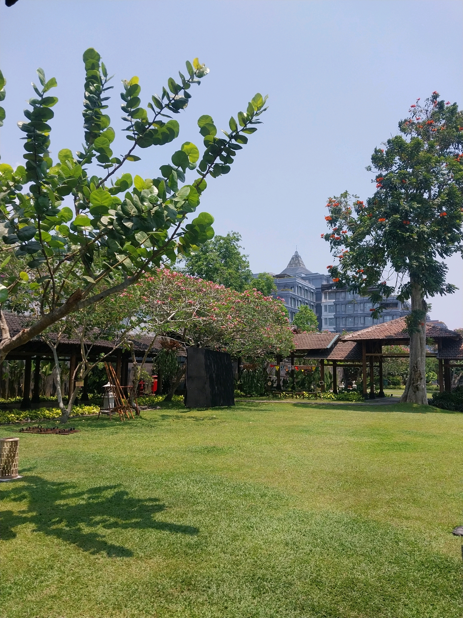 The image depicts an outdoor area with a well-maintained lawn in Ngaglik, Indonesia. In the foreground, a lush green lawn dominates the scene. To the left, a tree with large, round green leaves casts a shadow on the grass. Several wooden structures with tiled roofs are visible in the middle ground, resembling gazebos or pavilions. Behind these structures, a building with multiple stories and a distinctive architectural design, possibly a hotel or office building, stands against a clear blue sky. A tall tree with green leaves and scattered orange flowers is situated to the right of the frame, near one of the wooden structures. The overall ambiance suggests a serene park-like setting or resort.