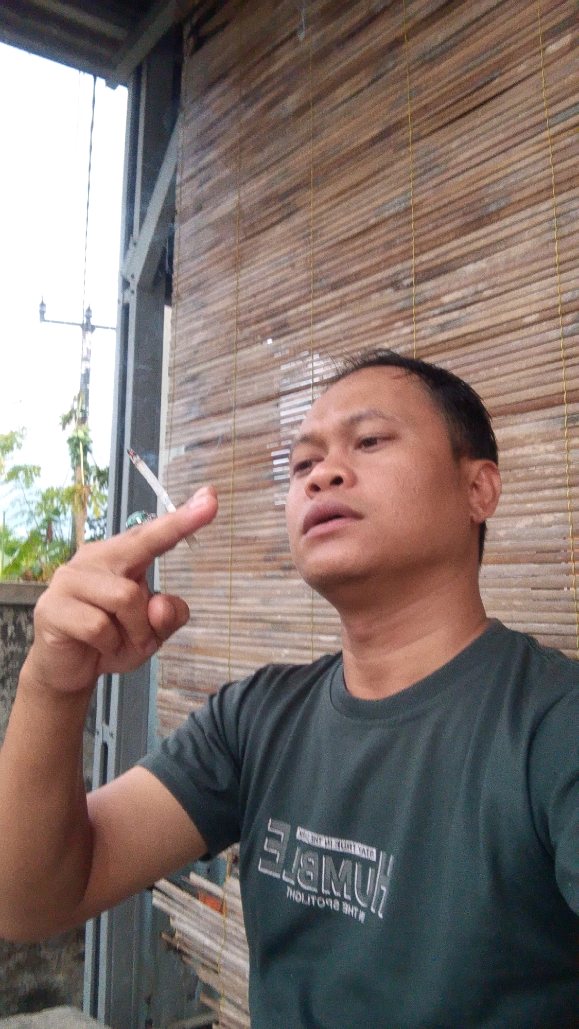 A man is taking a selfie, holding a lit cigarette between his index finger and thumb. He is wearing a dark green t-shirt with white text on it. The background consists of a wooden blind and some greenery visible through an opening. The scene appears to be outdoors, possibly on a porch or balcony, with some structural elements of a building visible. The lighting suggests it is daytime.