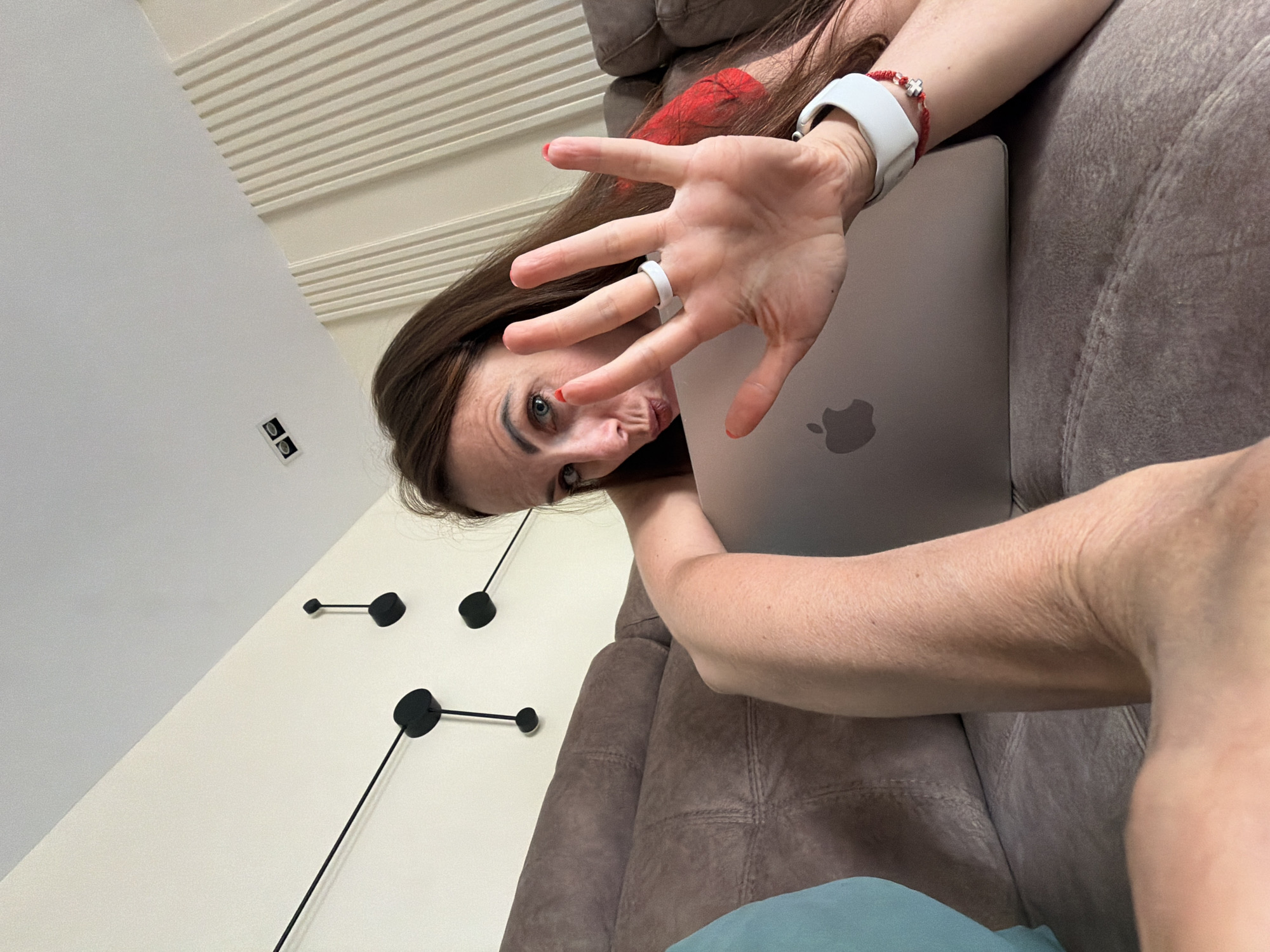 A woman is lying on a brown couch, holding a laptop. Her right hand is raised, palm facing the camera, with red nail polish visible on her fingernails. She is wearing a white ring on her left hand and a white watch with a red bracelet on her right wrist. A teal pillow is visible to the left of the couch. The wall behind her is white with a textured paneling on the right side. Black decorative wall fixtures are mounted on the wall. The scene appears to be indoors, possibly in a residential setting. There is no indication of the city of Birobidzhan, Russia in the image.