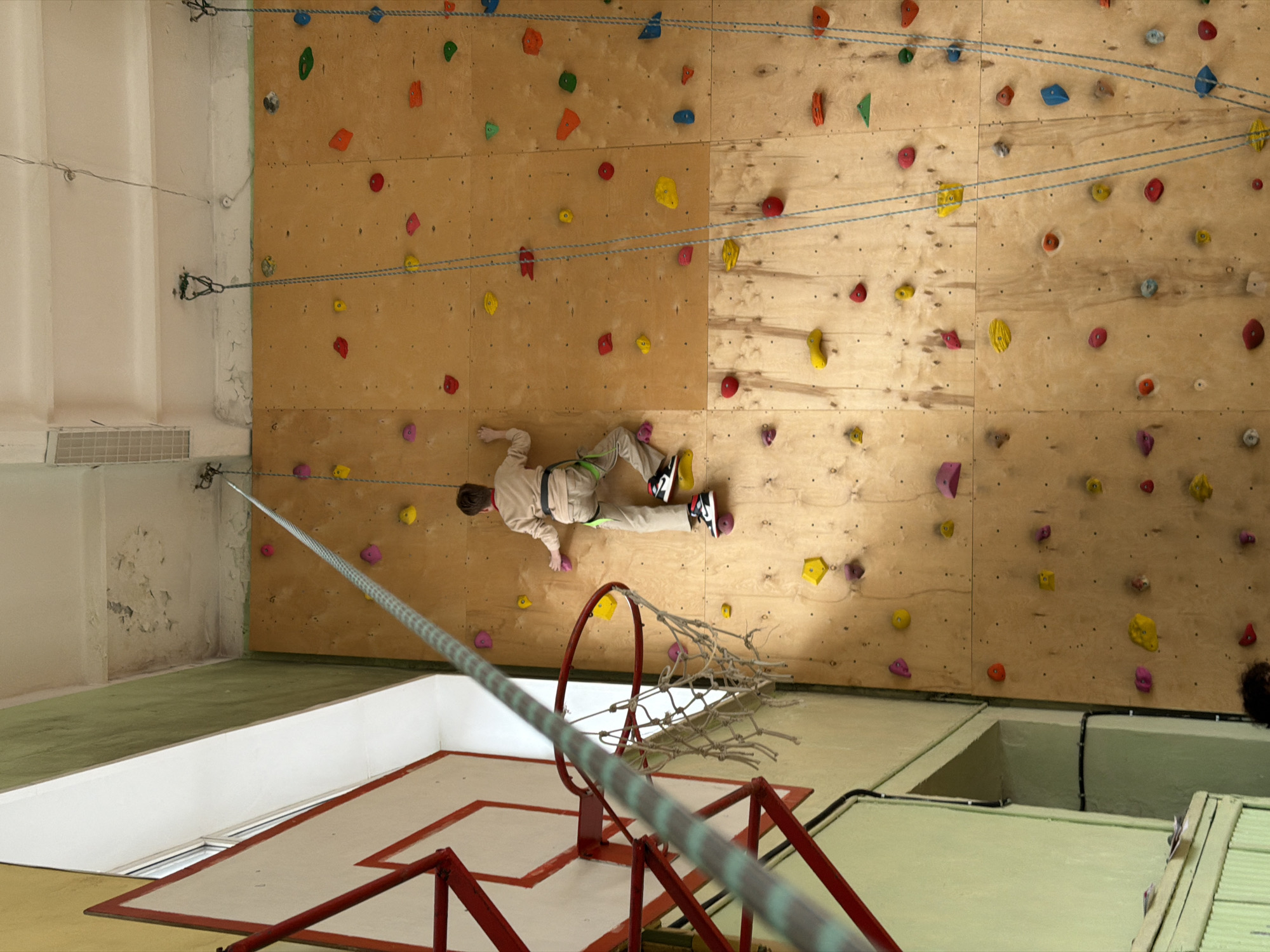 A wide-angle, interior shot depicts a climbing wall in what appears to be a gymnasium, possibly located in Izhevsk, Russia. A person in tan athletic gear is mid-climb on the wall, which is textured with colorful grips. The wall is paneled and has multiple climbing routes marked by blue ropes extending to the upper left. In the foreground, a basketball hoop is visible, with a net and red support structure. The floor is composed of alternating light green and white surfaces, indicating different sections of the gymnasium floor. The left side of the frame reveals a corner with peeling paint and an air conditioning unit.