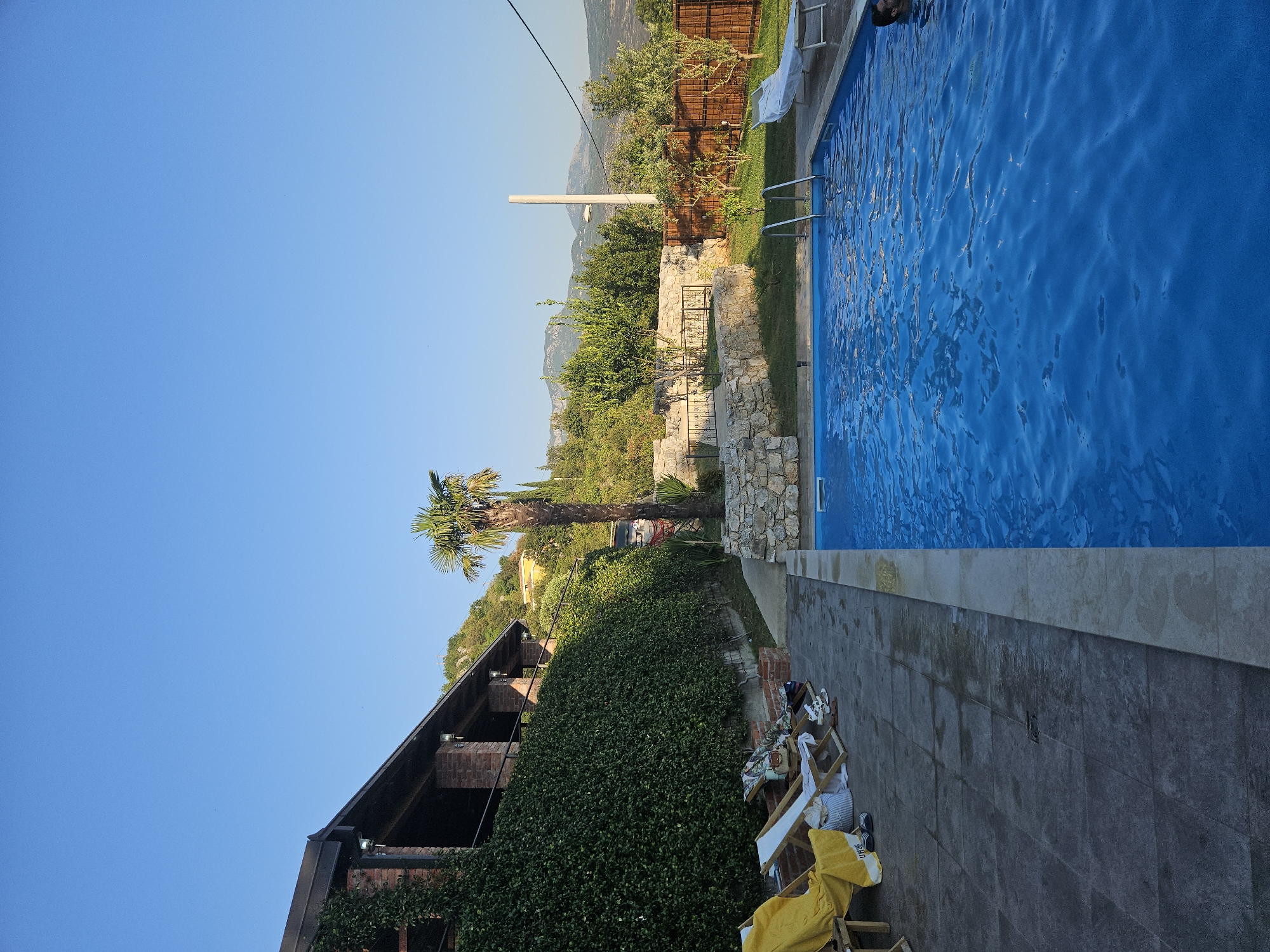 In an outdoor setting under a clear blue sky, a swimming pool with bright blue water takes up a significant portion of the right side of the image. A person's head is visible in the water on the far right. The pool has metal ladders for entry and exit. Adjacent to the pool is a tiled patio area with several lounge chairs. One chair on the left is draped with a yellow towel, and another nearby has a striped bag and sandals.
To the left of the patio, a building with a dark roof and brick accents is covered in dense green ivy. A tall palm tree stands near the building, with a stone retaining wall behind it. Beyond the pool and lounge area, a wooden fence encloses a grassy area with young trees. In the background, rolling hills covered in vegetation rise up, with mountains visible in the distance under the bright sky. The scene suggests a residential or resort property in a warm climate.