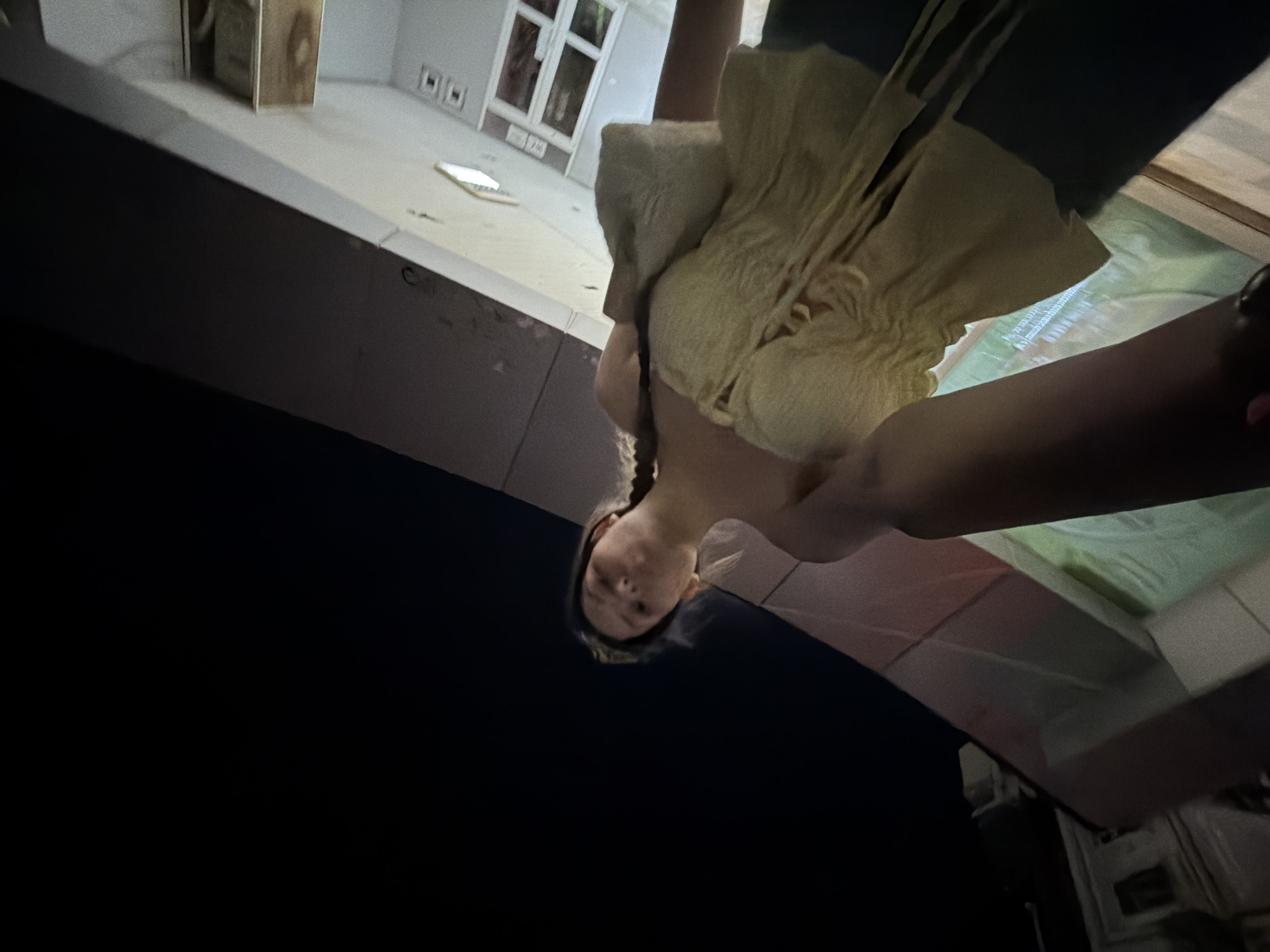 The image captures a female individual from an extreme low-angle perspective, looking directly down at the camera. She has light-colored hair and is wearing a textured, light-toned top, possibly beige or yellow. A dark strap, likely from a bag, is visible over her left shoulder. Her right arm is extended downwards, partially visible.
The setting is nocturnal, with a dark sky occupying the lower part of the frame, contrasting with the artificially lit background. The background reveals architectural details of a building, including light-colored walls and multi-paned windows. A wooden box or piece of furniture is partially visible, along with what appears to be a small, light-colored electronic device resting on a surface. Rectangular fixtures resembling electrical outlets are also present on a wall. Further into the background, there are hints of other structures.
Visible text, although blurry and indistinct, appears on a screen or printed surface in the background, showing characters resembling Hebrew script, including the word 'מחשבה' (machshava, meaning 'thought'). The photograph provides a unique, worm's-eye view of the subject against an urban backdrop, with the location context being Burgas, Bulgaria. The person's expression is neutral as she looks towards the lens.