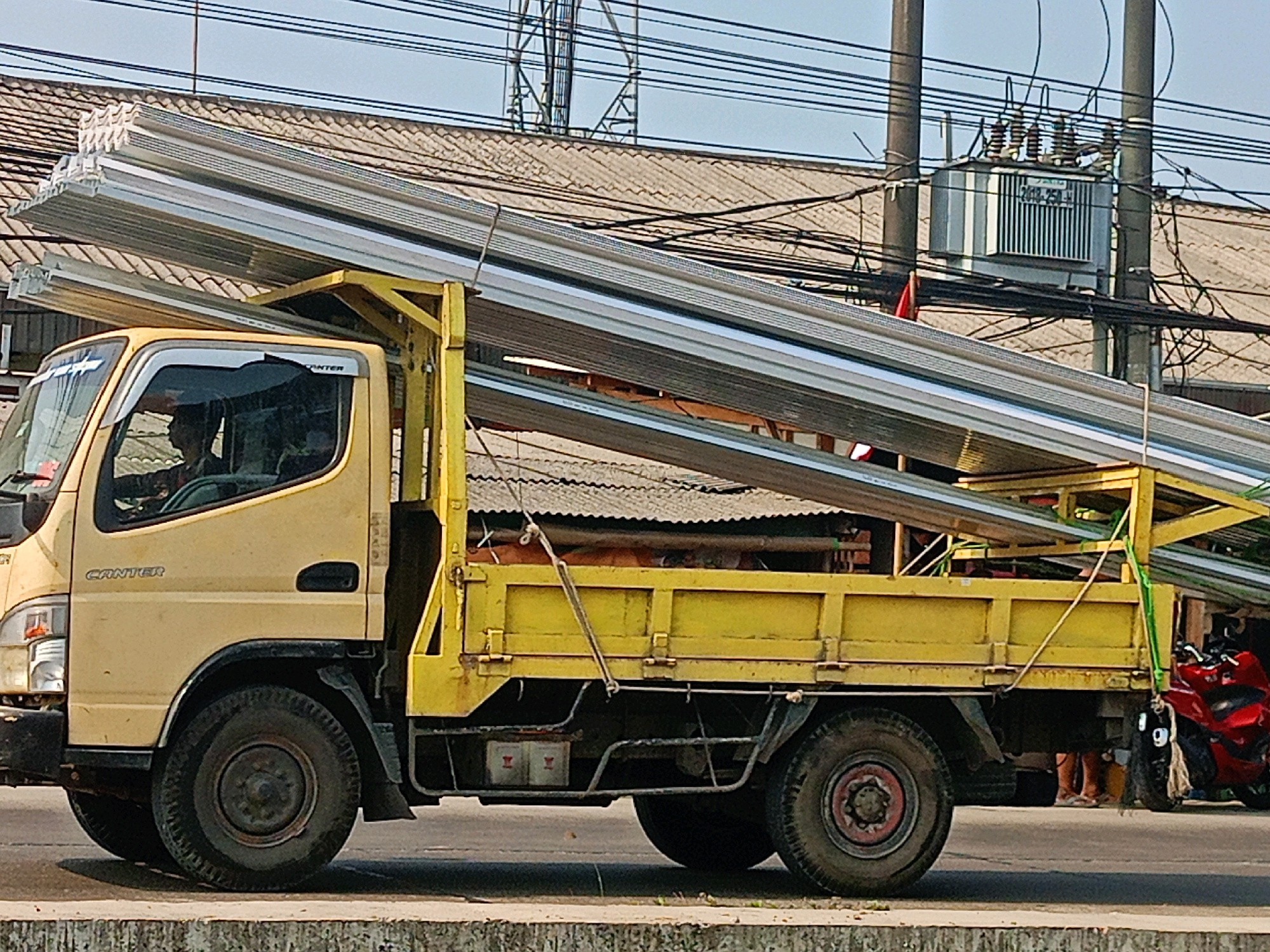 In Sriamur, Indonesia, a yellow flatbed truck, labeled "CANTER," is transporting a large load of long, slender, metallic objects, possibly roofing panels or construction beams, secured to its flatbed with yellow and green strapping. The truck's cabin is visible, showing a driver looking forward and another person inside. The truck is on a paved road, with a textured concrete barrier in the foreground. In the background, a tiled roof of a building and overhead power lines are visible, along with an electrical transformer on a utility pole. To the right of the truck, a red motorcycle is parked, and several people are standing near it. The scene appears to be outdoors, under a bright sky, suggesting daytime.