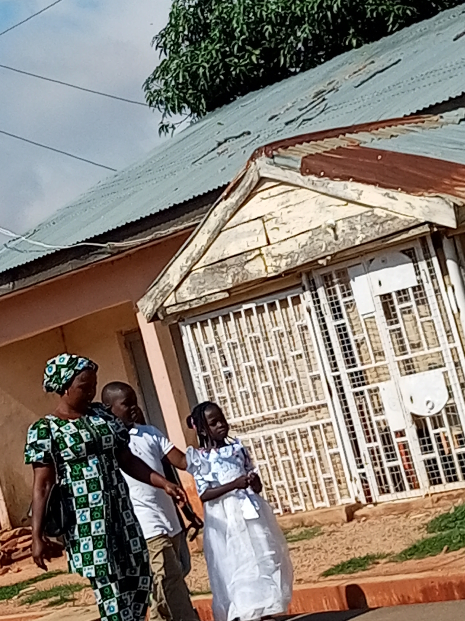 In the city of Unknown, Unknown, a group of three people, two women and one child, walk along a dirt path in front of a weathered building. The woman in the foreground wears a green and white patterned dress and a matching headscarf. She is followed by a man in a white shirt and khaki pants, and a young girl in a white dress. The building behind them has a corrugated metal roof and a white-painted facade with barred windows. A tree with green foliage is visible in the upper left corner of the image. The scene appears to be outdoors, during the daytime, with a cloudy sky.
