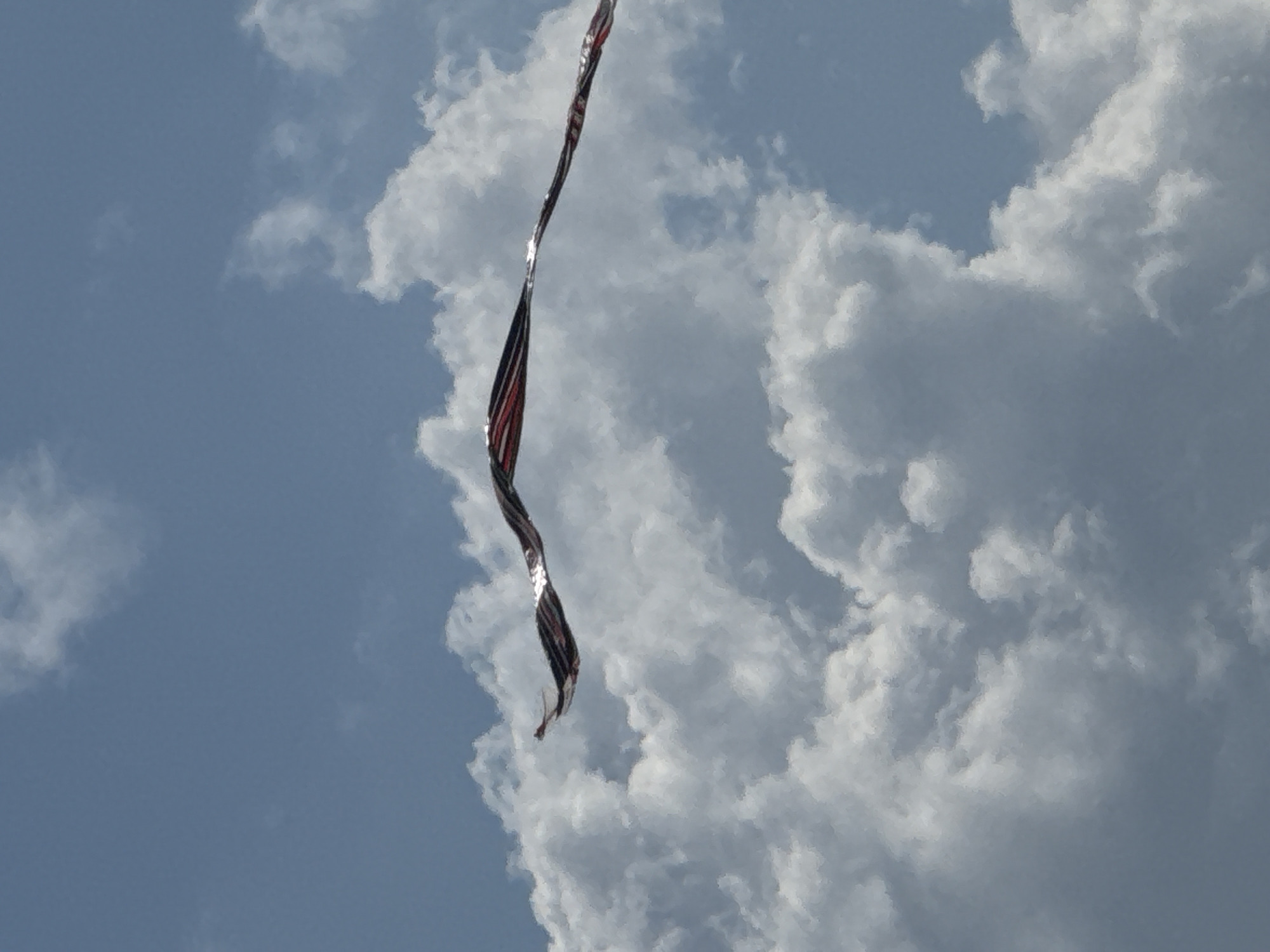 The image shows a ribbon-like object with red, white, and black stripes undulating against a backdrop of a cloudy blue sky. The object appears to be a kite tail or a decorative streamer. The clouds are large and puffy, suggesting a bright, possibly sunny day. The scene is devoid of any identifiable landmarks or human figures that would indicate a specific location in Denpasar, Indonesia.