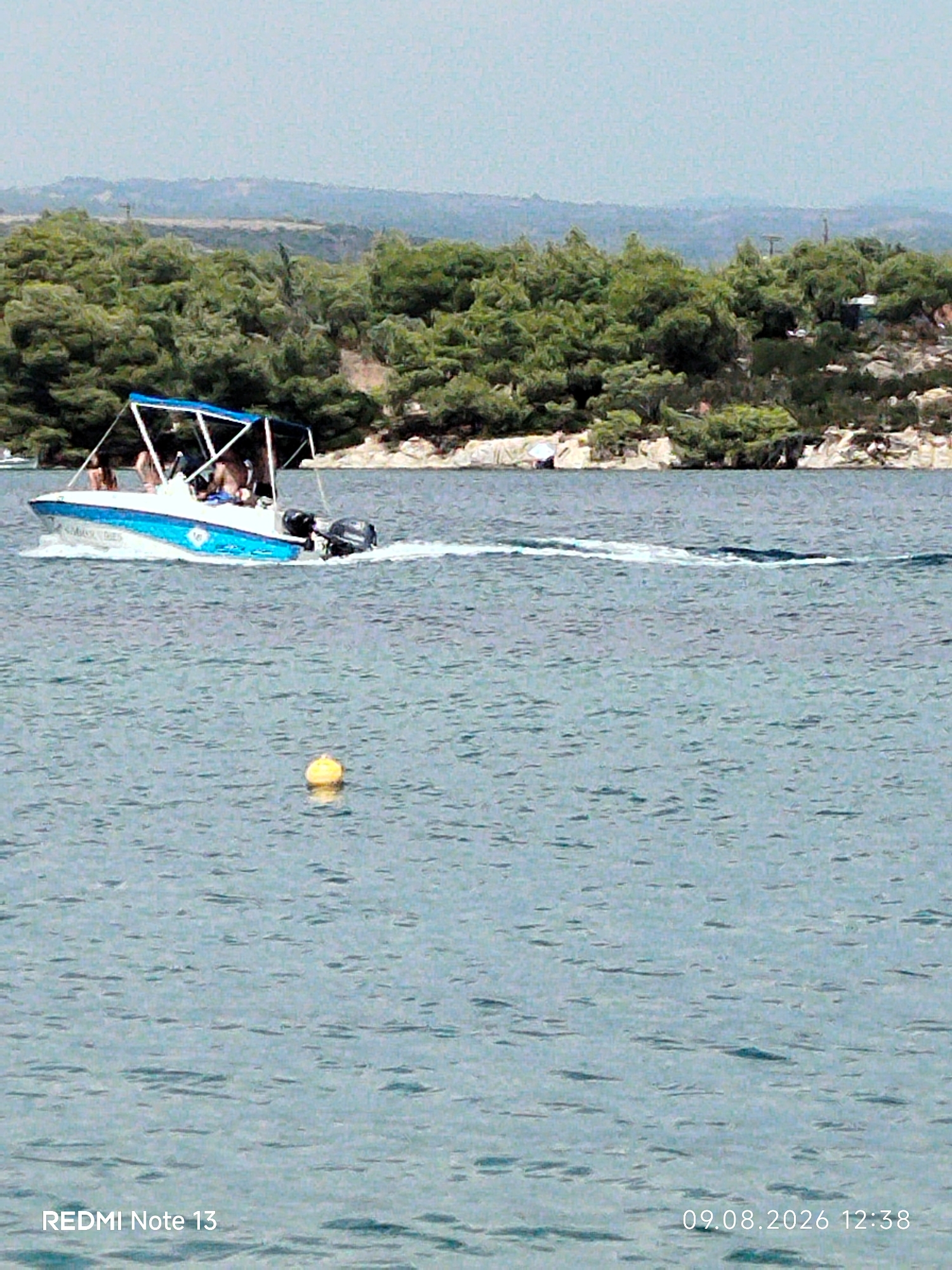 A white and blue speedboat with a bimini top and an outboard motor is moving across a body of water, leaving a wake behind it. Three people are visible on the boat, with two standing and one seated. The boat is heading towards the right side of the frame. In the foreground, a yellow buoy floats on the water. In the background, a lush green, tree-covered hillside with rocky outcrops rises from the water's edge, characteristic of coastal landscapes found in the Sithonia Municipal Unit of Greece. The sky is a light, hazy blue. The date and time "09.08.2026 12:38" and "REDMI Note 13" are overlaid on the bottom right and bottom left corners of the image, respectively.