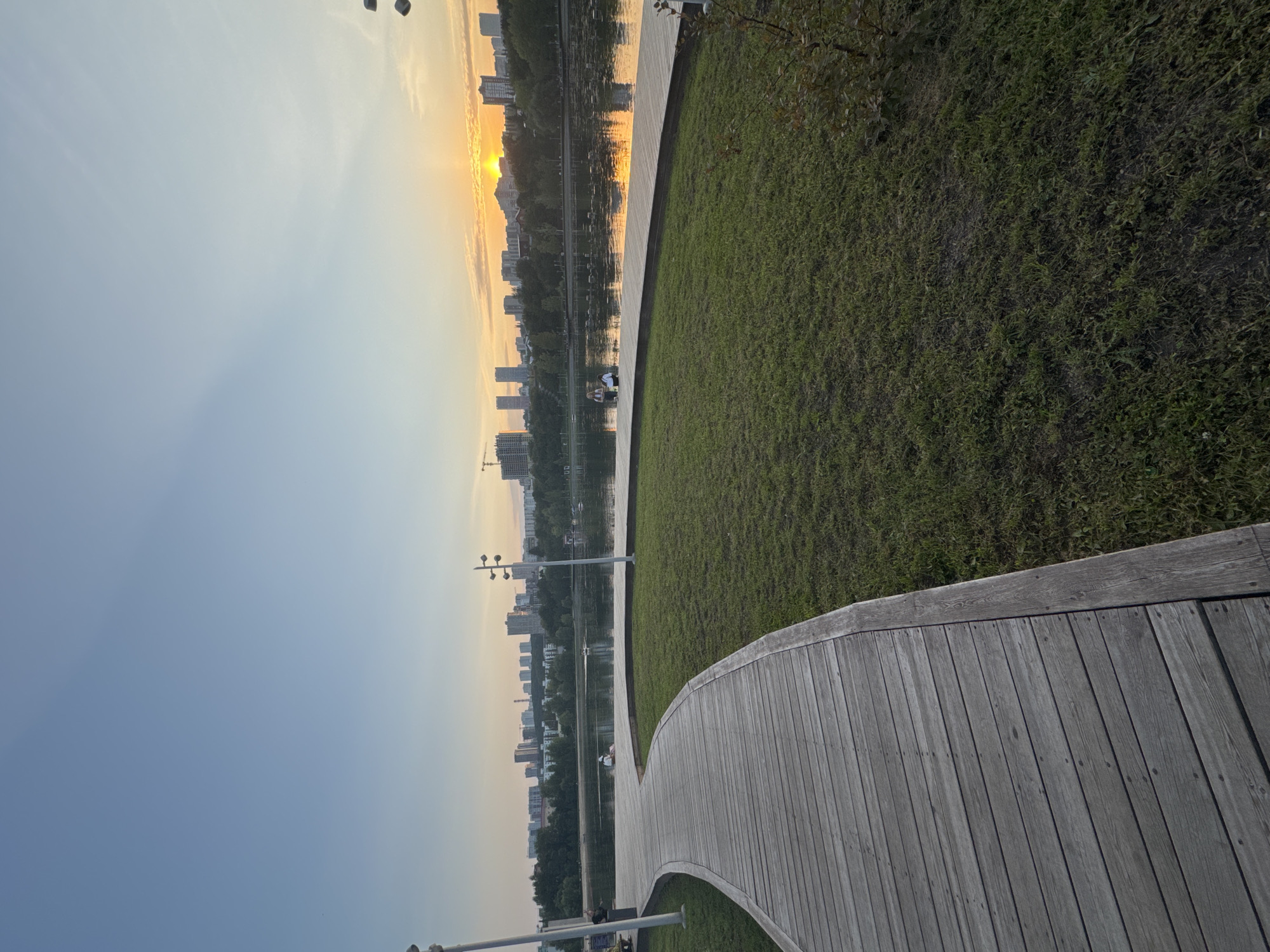The image depicts a sunset scene at a park in Ufa, Russia. A wooden walkway curves through a grassy area, leading towards a calm lake. In the distance, a city skyline with numerous buildings is visible. The sun, partially obscured by clouds, is setting over the horizon, casting a warm, orange glow on the sky and water. Several boats are scattered on the lake, and a few individuals can be seen near the water's edge. The overall atmosphere is serene and peaceful.