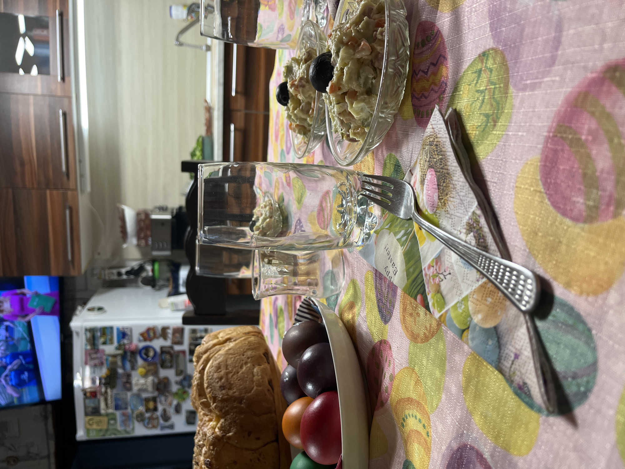 The image depicts a table setting with food. There are bowls of salad with olives, empty glasses, a loaf of bread, and a bowl of colored eggs on a tablecloth with Easter egg patterns. Cutlery, including a fork and a knife, is also visible. The setting is indoors, likely a home. In the background, a refrigerator covered in magnets and a television screen are visible. It is impossible to know if this scene is in the city of Pobeda, Bulgaria without additional information.