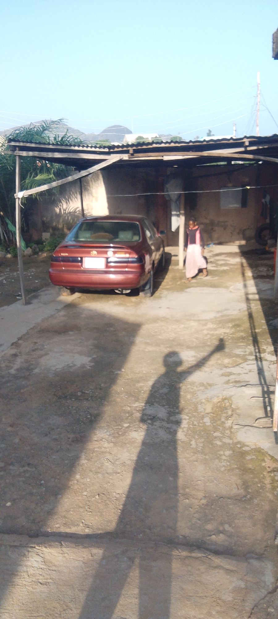 A maroon sedan is parked in a dirt lot beneath an open-sided shelter. The shelter has a corrugated metal roof and wooden support beams. To the right of the car, a young girl in a pink and black outfit is standing. A long shadow of a person with an outstretched arm is cast on the ground in the foreground. In the background, there are some palm trees and distant hills under a clear blue sky.