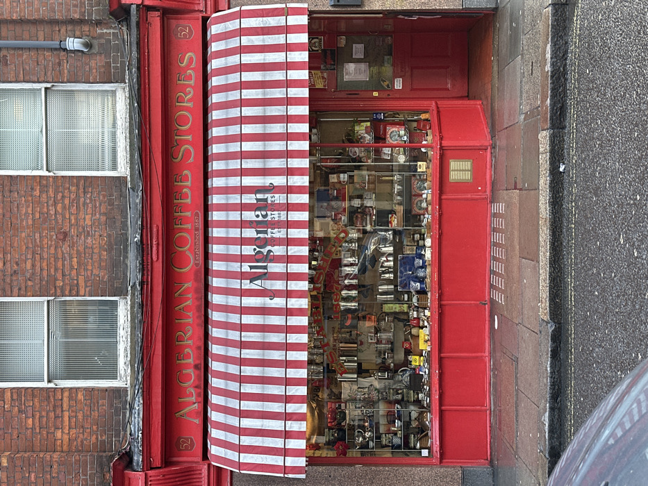 This image presents the exterior of the Algerian Coffee Stores, located at 52 Old Compton Street in Soho, London, United Kingdom. The building's upper facade is red brick, featuring two windows with white frames and textured privacy glass. Below this, a bright red fascia displays "ALGERIAN COFFEE STORES Established 1887" in gold lettering, with the number "52" at each end. A red and white striped retractable awning, also branded "Algerian Coffee Stores Est. 1887", extends over the ground-level shop front.
The ground floor features a large, glass display window and a recessed doorway, both painted in a vibrant red. The shop window is densely packed with merchandise, including numerous metallic coffee makers (moka pots, French presses), grinders, cups, and packaged goods arranged on internal shelves. Reflections of the street and sky are visible on the glass. The entrance consists of a red wooden door, with several notices or papers affixed to its surface. A dark grey letterbox is set into the stone wall immediately to the right of the shop front.
The pavement in front of the store is wet and grey, with tactile paving strips visible near the entrance. A dark curb separates the pavement from a wet, asphalt road. The front section of a dark-colored vehicle is visible in the lower-left corner of the image, parked on the street. No individuals are present in the scene, and no overt actions or interactions are depicted.