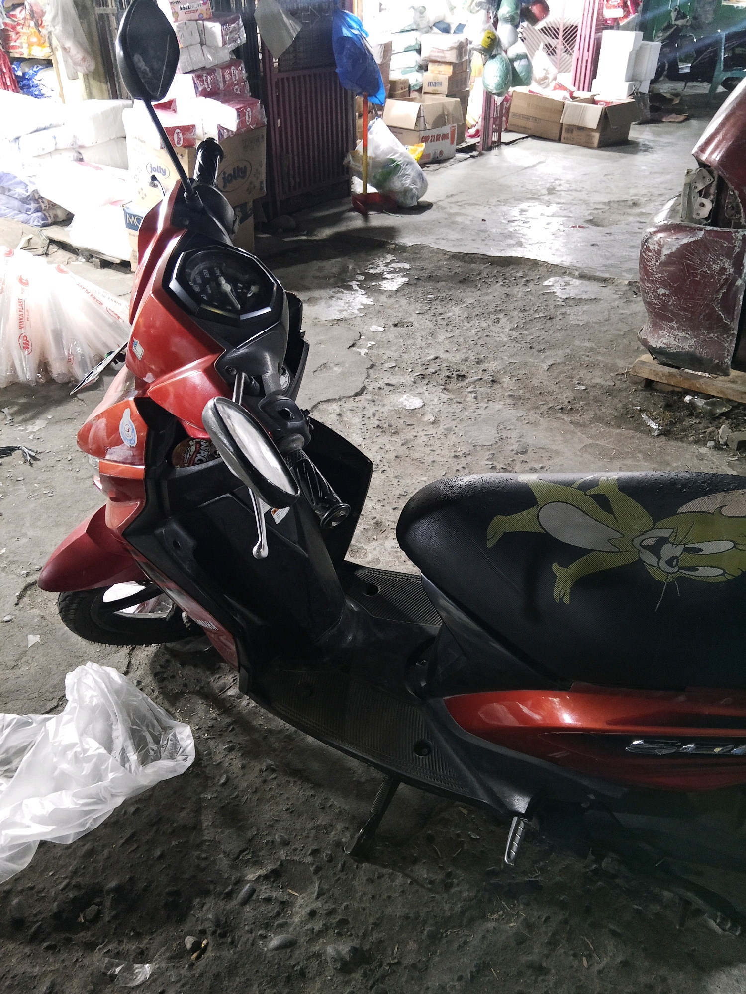 The image shows a red and black scooter parked on a dirt and concrete ground. The scooter has a seat with a cartoon character print. In the background, there are stacks of boxes and some plastic bags. The scene appears to be an outdoor area in a city, possibly a marketplace or storage area. The lighting suggests it might be late evening or an indoor space with artificial lighting. No people are visible in the image. The environment is cluttered with various objects, indicating a busy or storage-focused location.