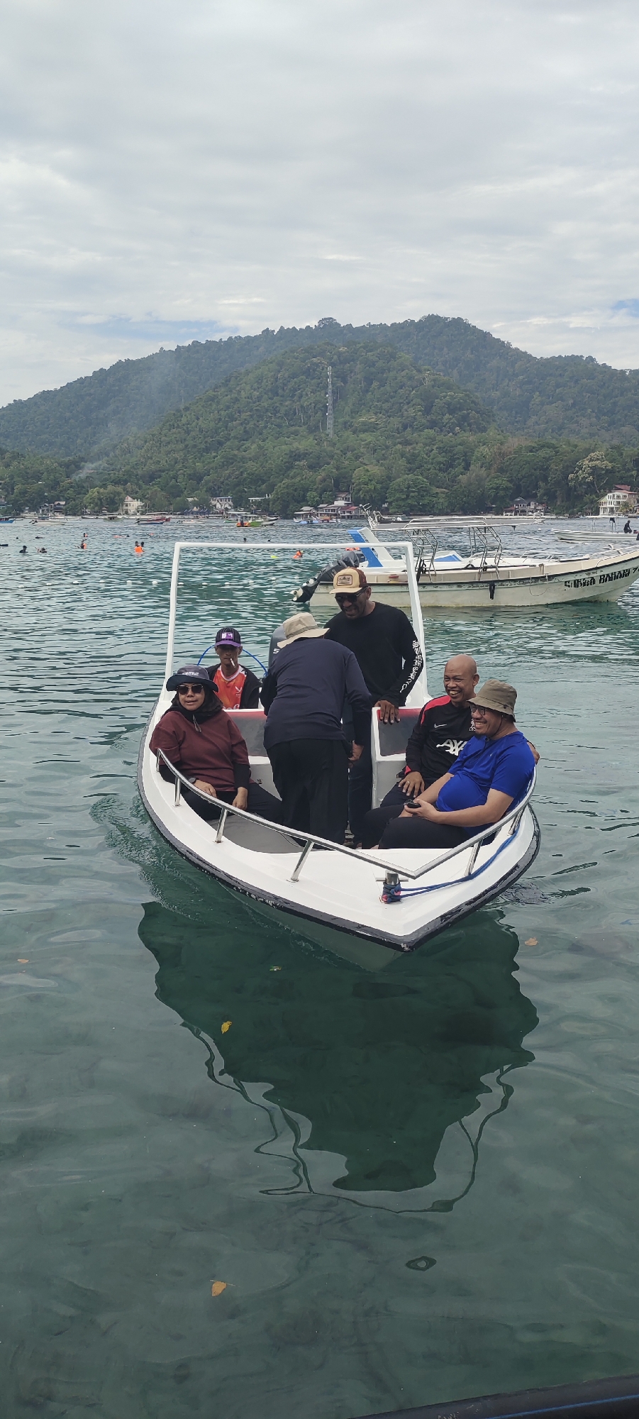 A group of six people are in a small white boat on the water, with a lush, green, mountainous island in the background. The sky is overcast. The boat is a narrow, pointed vessel with a white frame structure on top, and it appears to be moving slowly.
In the boat, from left to right:
- A woman wearing a maroon hoodie and a dark headscarf is seated on the left side.
- A young man in an orange shirt and a purple baseball cap is standing behind her, holding a cigarette.
- A man in a black long-sleeved shirt and a baseball cap is standing and facing towards the right.
- A man in a dark blue shirt and a bucket hat is seated on the right side, with his arm around the man next to him.
- A man in a red and black shirt, with a bright red collar, is seated in the middle. He is laughing and looking towards the right.
- Partially visible behind him is another person in a black shirt.
The water is clear and green, with reflections of the boat and the people visible. There are other boats and swimmers in the distance, and the shore of the island has buildings and trees. The mountainous island suggests a tropical location. This scene is likely in the waters around Sabang, Indonesia, a popular tourist destination known for its natural beauty and water activities.