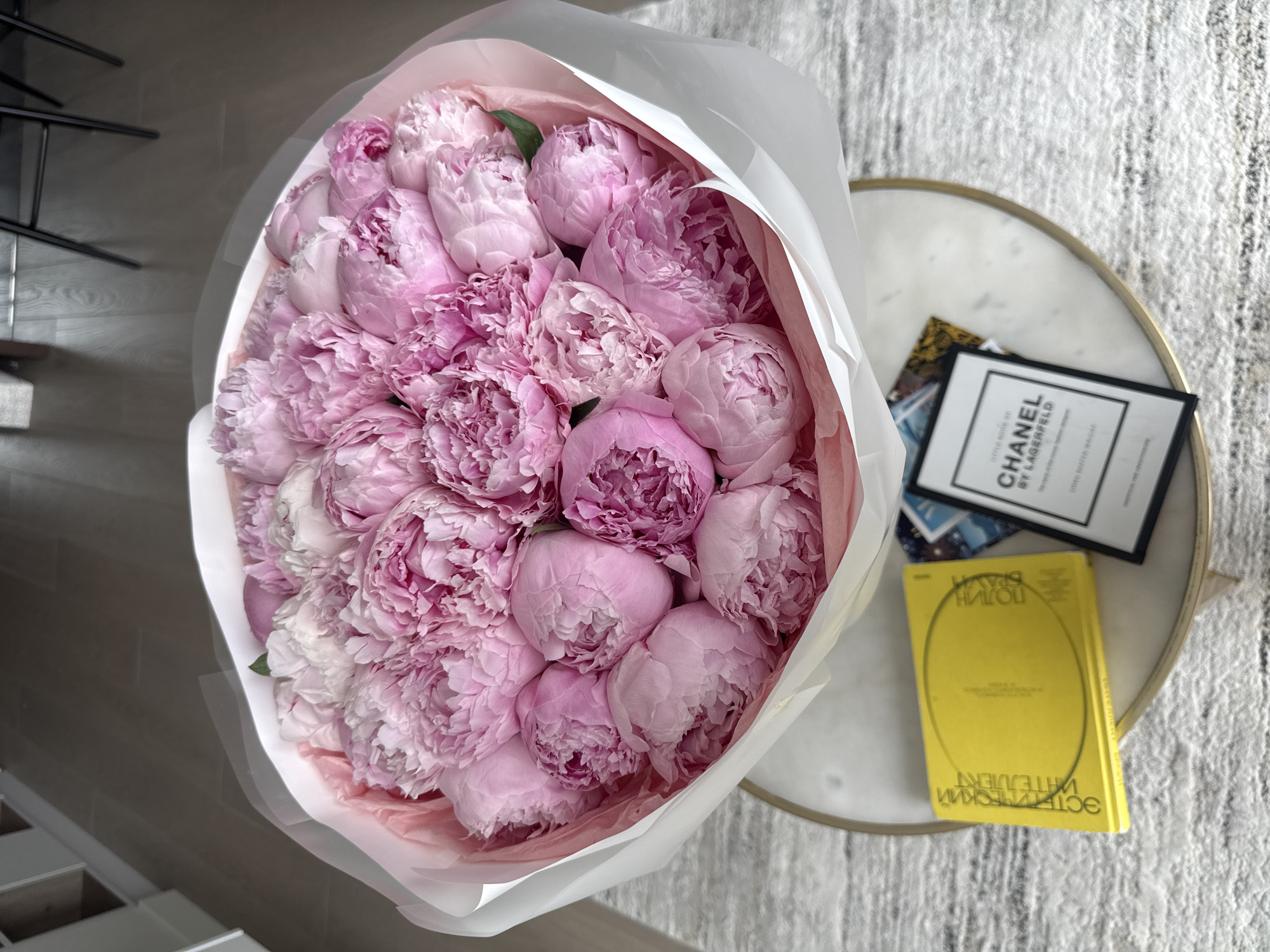 This media file captures a top-down view of a large, lush bouquet of pink peonies, presented in an indoor setting in Khimki, Russia. The bouquet, the primary subject, consists of numerous light to medium pink peony buds and partially opened flowers, showcasing varying shades and textures of the petals. It is wrapped in soft pink tissue paper, further encased by a layer of white opaque paper, creating a voluminous and elegant presentation.
The bouquet is positioned on or next to a round side table featuring a light-colored surface, possibly marble or a marble-effect material, framed by a thin gold-colored rim. On the table, to the right of the flowers, are two books. One is a black and white framed book titled "CHANEL BY LAGERFELD." The other is a smaller, vibrant yellow book with Cyrillic text. The visible Cyrillic text appears to be "ЭСТЕТИЧЕСКИЙ МАТЕРИАЛИЗМ" (Esthetic Materialism) and "ПРАВОЙ ПОЛЮС" (Right Pole), though it is partially mirrored and slightly blurry.
The table and flowers rest on a textured, light-colored rug with a white and grey patterned design. In the blurred background on the left, a dark, abstract metallic structure, possibly part of a piece of furniture, is visible, with a light wooden floor beneath it.
No specific activity is taking place; the image is a composed still life. The peonies appear fresh, with many buds still tightly closed, suggesting they are newly acquired. The overall impression is one of modern elegance and a stylish, serene environment. The time of day and weather are not discernible from the indoor lighting.