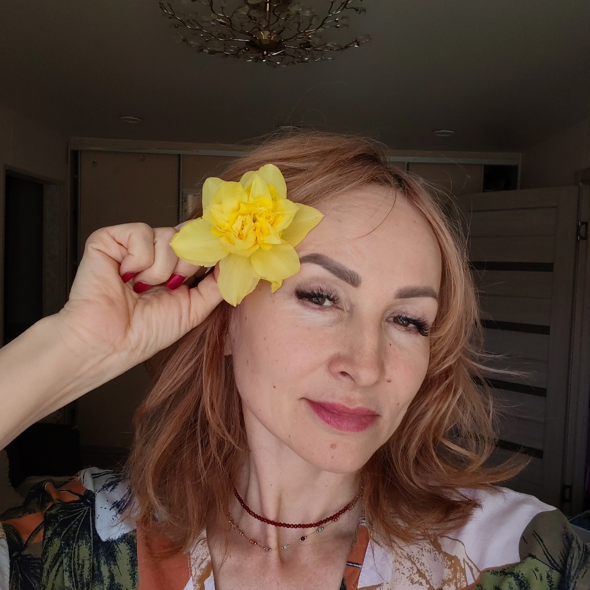 A woman poses indoors, holding a yellow flower near her head. The background shows a room with a chandelier, doors, and a closet. The location is the city of Chaikovsky, Russia.
