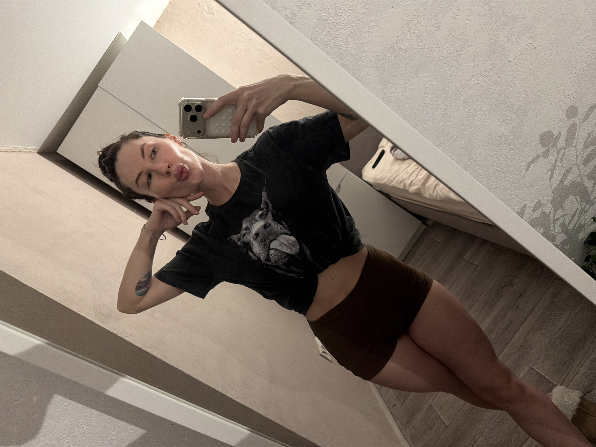 A woman is taking a selfie in a mirror. She is wearing a dark t-shirt and brown shorts. Her hair is pulled back, and she is making a kissing face. The room appears to be a bedroom, with a bed visible in the background. A plant casts a shadow on the wall. The location is potentially a residence in the city of Perm, Russia.