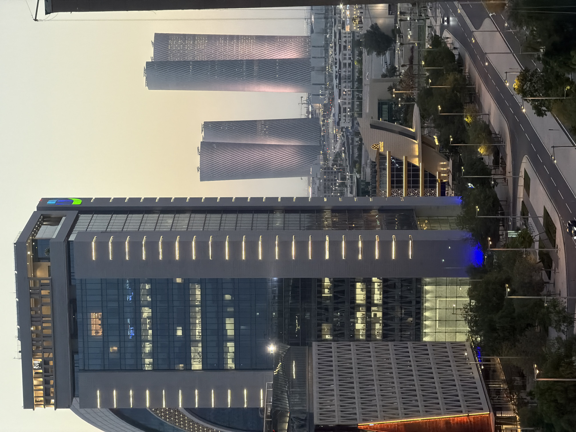 This image captures a cityscape at dusk, featuring modern architectural elements and a hazy sky. In the foreground, a tall, rectangular skyscraper dominates the left side of the frame, its facade adorned with horizontal illuminated strips and a grid of windows revealing interior lighting. To the left of this building, a curved structure with a patterned texture is partially visible. Adjacent to the main skyscraper, another building with a patterned facade and illuminated edges stands. Below these structures, a street curves through the scene, lined with trees and lampposts. Several cars are visible on the road. In the mid-ground, a distinctive building with a sloped roof and illuminated signage is present, with more urban development and greenery behind it. In the background, three tall, cylindrical towers with ribbed facades are illuminated by a soft, pinkish light. The sky is a pale yellow or orange, indicating either sunrise or sunset. The overall impression is one of a contemporary urban environment.