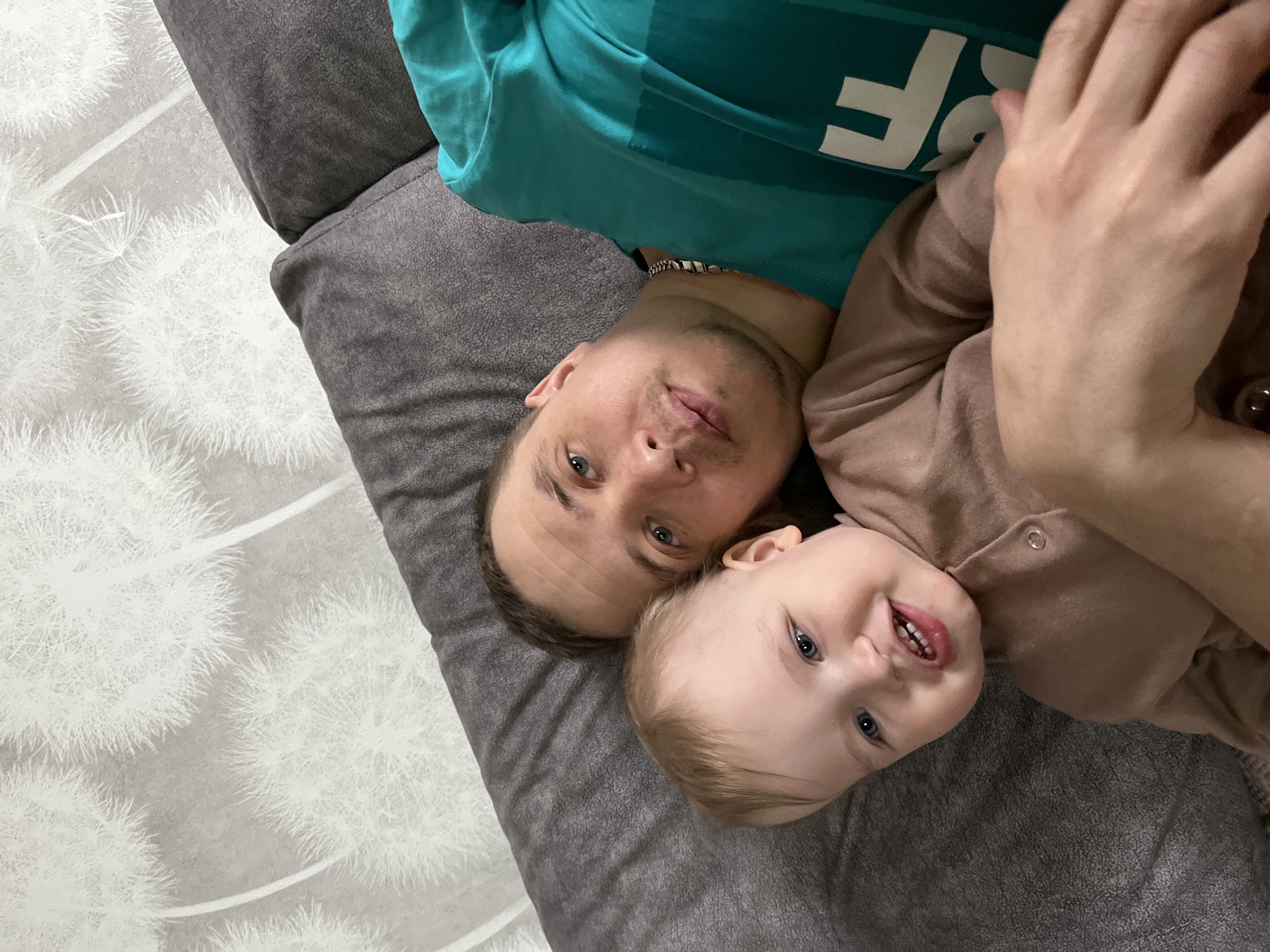 An adult male and a young child are positioned in a close-up, eye-level photograph within an indoor residential setting in Chaikovsky, Russia. Both individuals are lying on a dark gray, upholstered surface, consistent with a sofa or bed. The adult male, with short dark hair, light stubble, and blue eyes, wears a teal long-sleeved shirt with white lettering on the left sleeve and a silver-colored chain around his neck. He is positioned to the right, looking directly at the camera with a neutral expression. The young child, with light blonde hair and blue eyes, is situated to the left of the male, smiling towards the camera and showing several lower teeth. The child wears a light brown, long-sleeved, collared one-piece outfit with light-colored buttons down the front. The adult male's right hand is visible, resting on the child's lower body. The background features a light gray wall with a repeating pattern of large white dandelion seed heads.