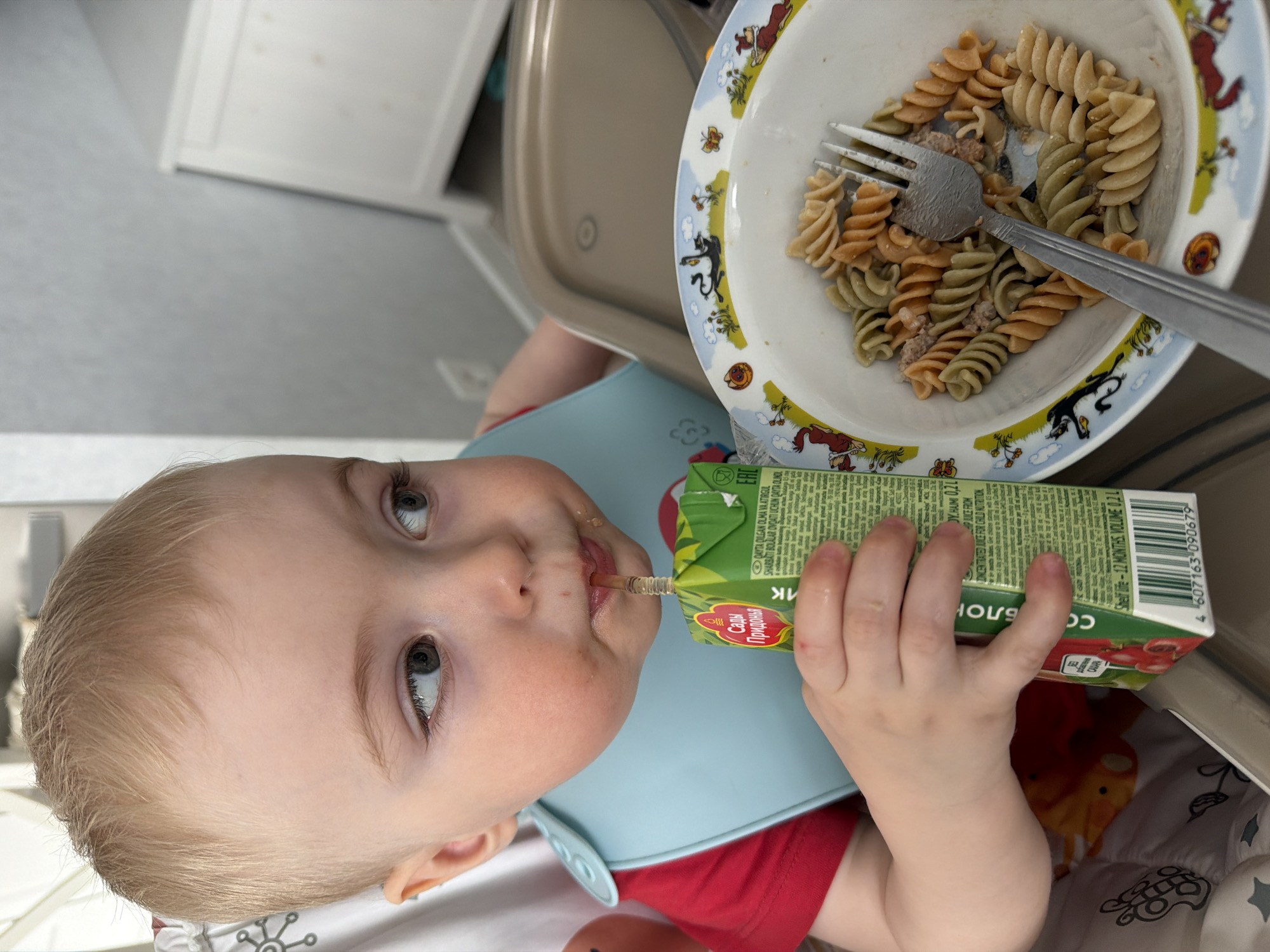 A close-up, slightly angled overhead shot shows a young child in a highchair drinking from a juice box. The child has light skin, blonde hair, and blue-grey eyes. A light blue bib covers a red shirt. The juice box is green and red with text in a non-English language. The child's hand holds the juice box. To the child's upper right is a plate of multicolored rotini pasta with a silver fork. The edge of the plate is decorated with cartoon animal illustrations. The background includes a white cabinet, a beige highchair, and a patterned seat cover. The juice box identifies the product as "Сады Придонья" (Sady Pridonya), a brand that is manufactured in Russia. No location of Votkinsk, Russia is discernible in the image.