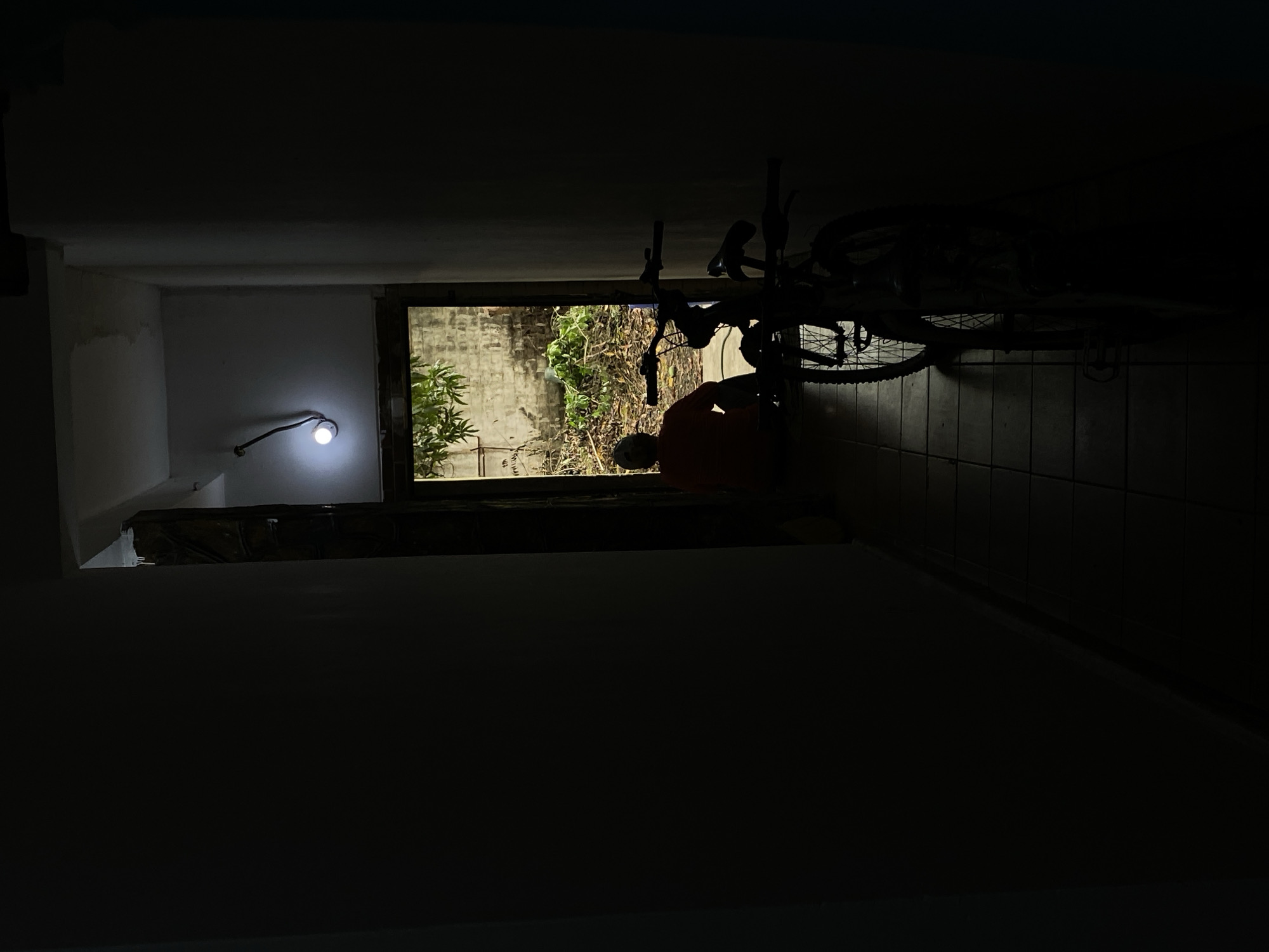 In a dimly lit, narrow indoor walkway with tiled flooring, a silhouette of a bicycle stands prominently in the foreground. The walkway opens to an outdoor area where a person, also in silhouette, is seated facing away from the camera. Behind the seated figure, a brick wall is visible, overgrown with vegetation. A single light bulb hangs from the ceiling, illuminating the scene with a faint glow. The image conveys a sense of solitude and stillness, characteristic of a quiet moment captured in a residential area of Tinaquillo, Venezuela.