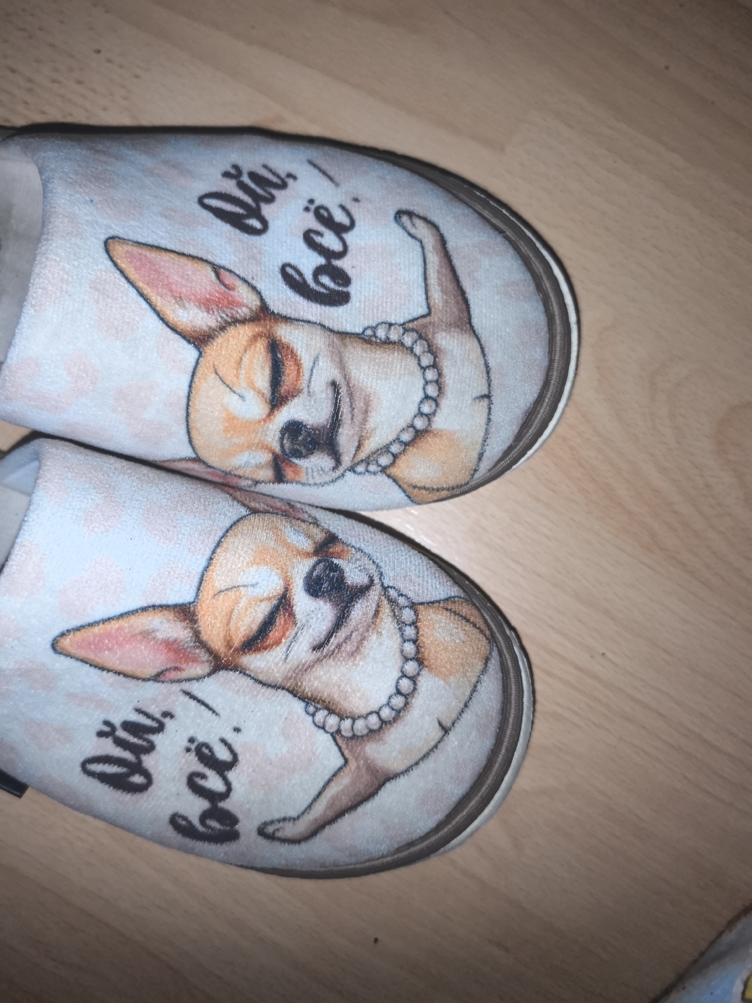 A photograph shows a pair of indoor slippers resting on a wooden surface. The slippers feature a light blue fabric with a pattern of small, scattered pink dots. Each slipper is decorated with an image of a stylized chihuahua wearing a pearl necklace. The dog is depicted with closed eyes and a serene expression. To the left of the dog image on each slipper, there are black Cyrillic words that read "Ой, всё!". The soles of the slippers are a darker shade, likely rubber or a similar material for grip. The wooden surface beneath the slippers appears to be a light brown color with visible grain. The slippers are positioned next to each other. There are no people visible in the frame. The slippers may be located in Moscow, Russia.
