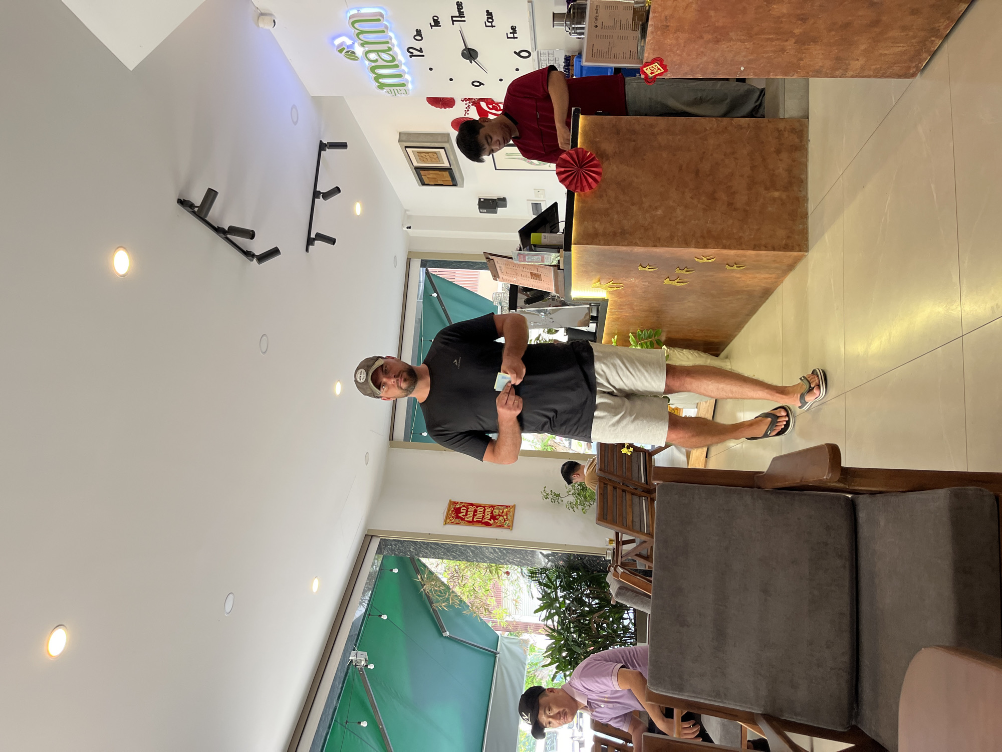 The image depicts the interior of a cafe, likely in Nam Nha Trang Ward, Vietnam. The cafe features a modern, clean aesthetic with light-colored tiled flooring and a counter area.
In the foreground, a man wearing a black t-shirt, light-colored shorts, and a baseball cap stands in the center of the frame, holding what appears to be cash in his hands. He is looking towards the camera. To his left, a man wearing a purple polo shirt and a dark cap is seated at a table, looking towards the man in the center.
The counter area, to the right of the man in the center, is manned by an employee wearing a red polo shirt and dark pants. He is interacting with something on the counter, possibly a register or making a drink. Behind the counter, a clock with Vietnamese numerals is visible on the wall, alongside a neon sign that reads "mam cafe." Various decorative elements, such as red paper fans and gold decals on the counter, add to the cafe's ambiance.
The background reveals more seating areas and large windows that allow natural light to enter the space, revealing greenery and a street scene outside. The overall atmosphere suggests a casual and welcoming environment.