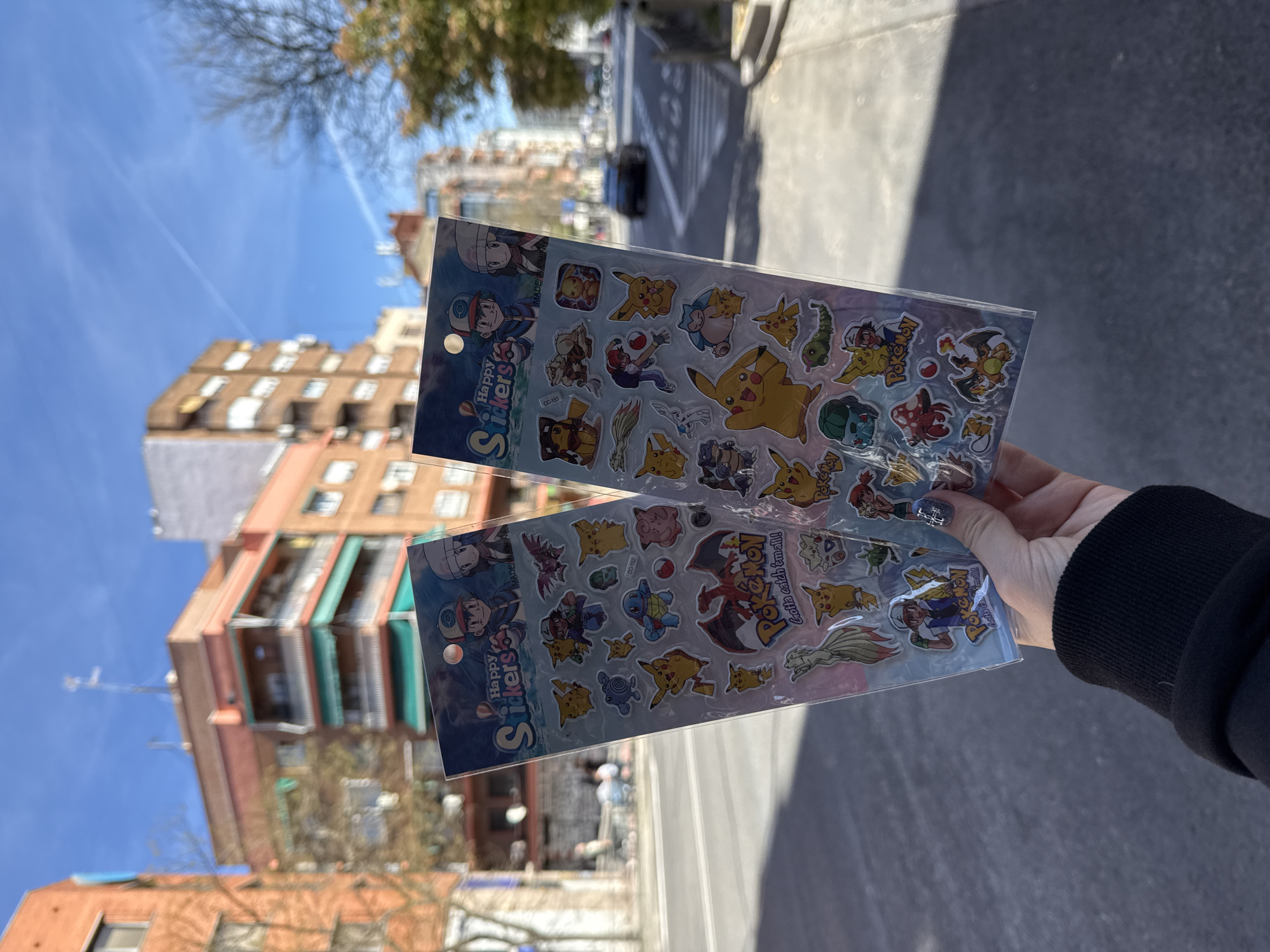 A medium shot captures a person holding two sheets of Pokemon stickers against an urban backdrop. The stickers, encased in clear plastic, feature various Pokemon characters such as Pikachu, Charizard, and others. The packaging prominently displays the word "Pokemon" and the slogan "Gotta catch 'em all!" The person's hand and part of their arm, clad in a black jacket, are visible.
In the background, an urban street scene unfolds, suggesting the location is Madrid, Spain. Architectural elements include a multi-story building with balconies, typical of Madrid's residential areas. The street appears to be paved and includes pedestrian crossings. A car is faintly visible in the distance, adding to the sense of urban activity. The sky above is bright blue, indicating clear weather conditions. The scene is generally sunny, casting shadows and enhancing the visibility of the objects and surroundings.